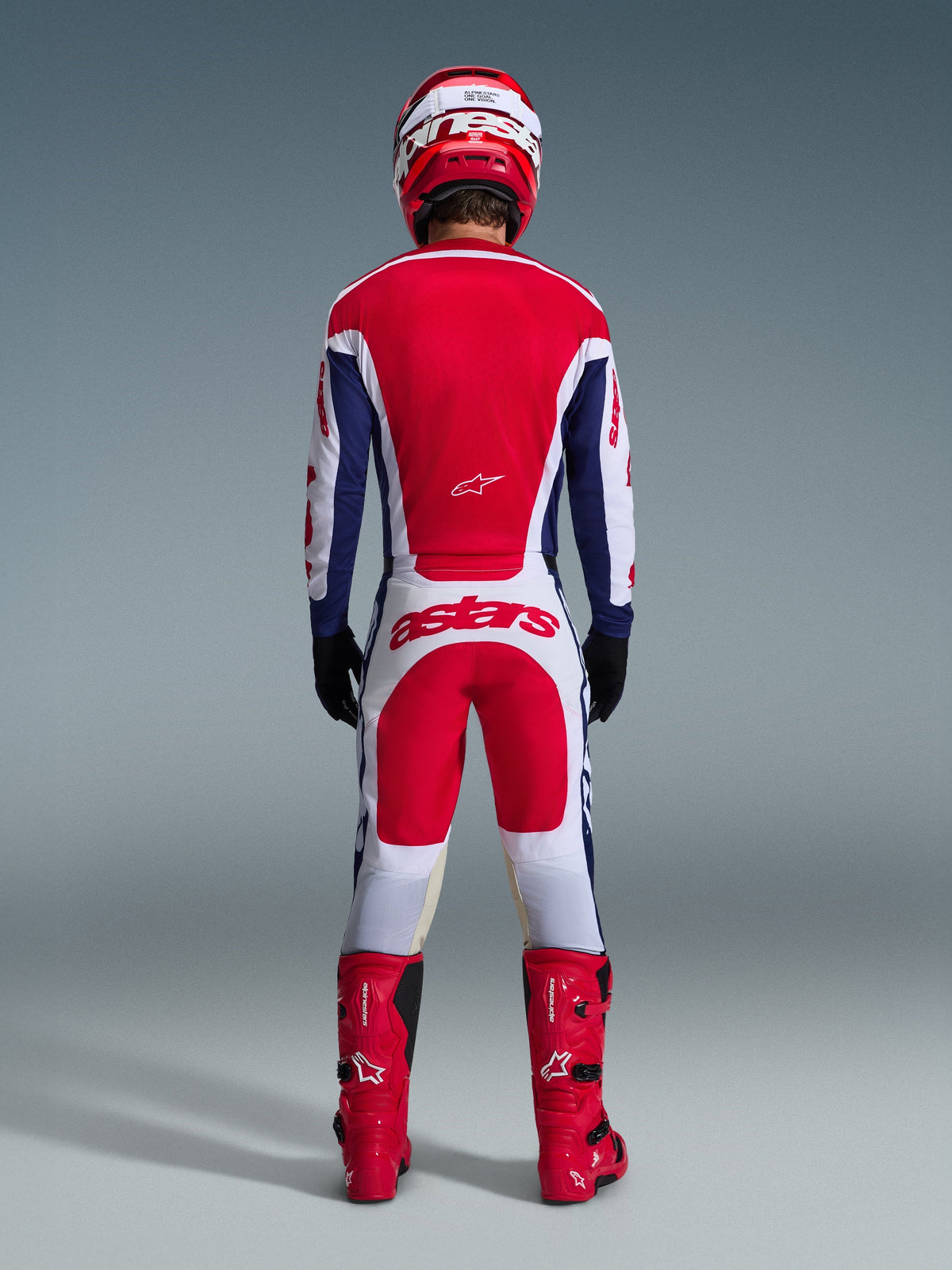 Motocross rider viewed from behind wearing the 2026 Racer Riway Gear Set, Red and blue jersey with white accents, white pants with red panels and 'astars' logo on the seat, coordinated with a red helmet, red boots, and black gloves, high-performance racing gear for off-road
