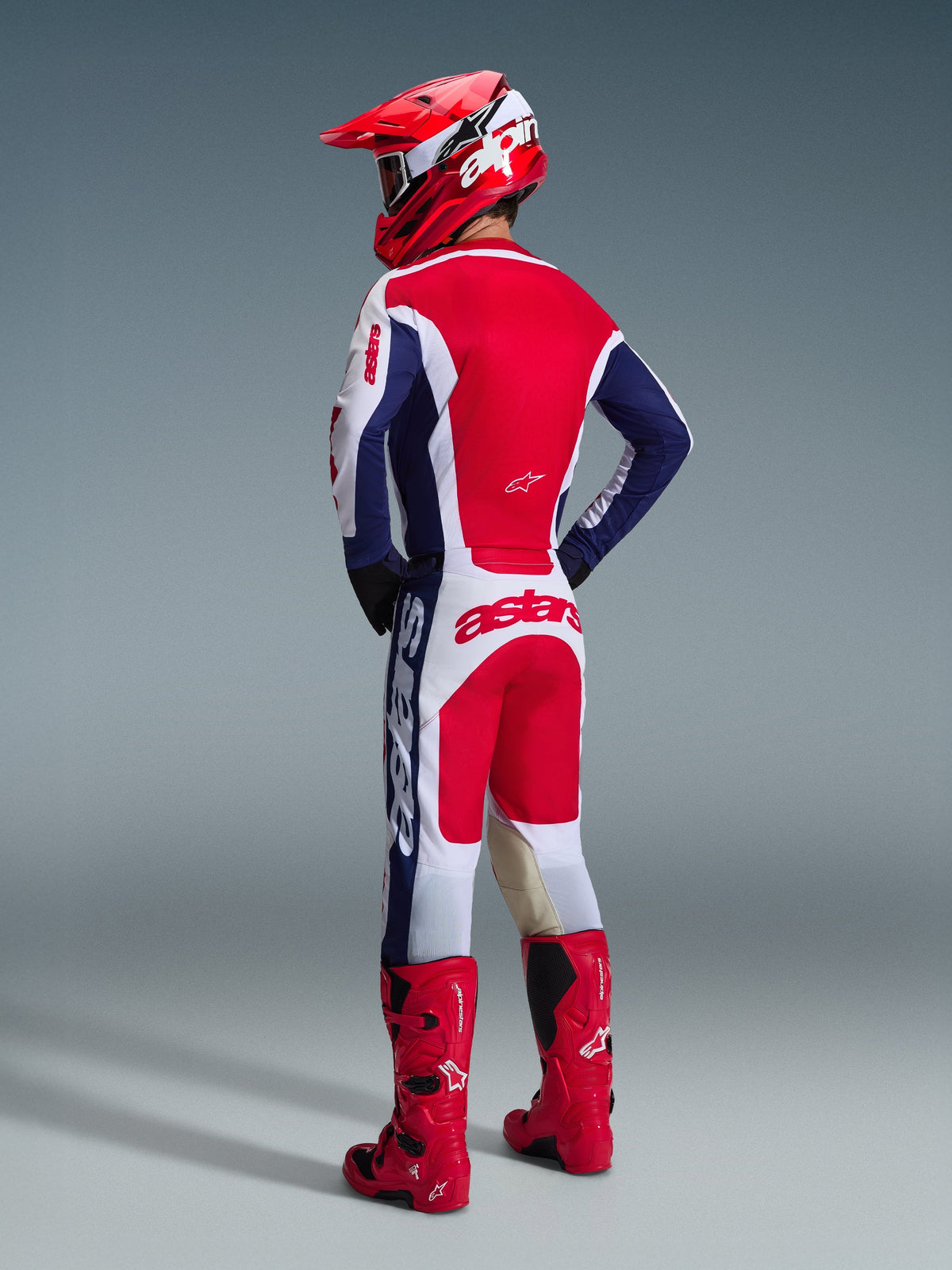 Motocross rider seen from behind wearing the Alpinestars 2026 Racer Riway Gear Set in White Blue Red, including a long-sleeve jersey with Astars branding and matching pants, coordinated with a red helmet and red off-road boots.