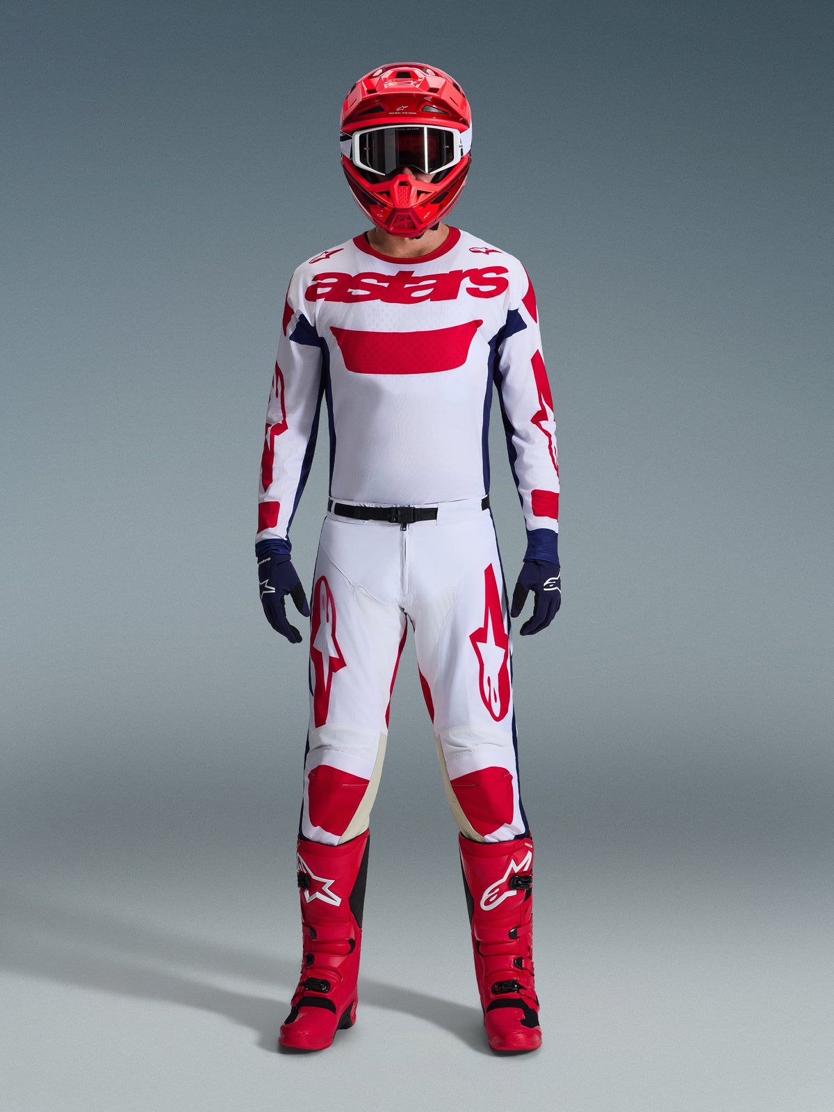 Pilote de motocross portant un équipement de course complet, maillot Alpinestars Racer Riway 2026 blanc bleu rouge, pantalon assorti blanc et rouge, casque rouge, bottes rouges et gants bleu marine, tenue coordonnée pour la course tout-terrain