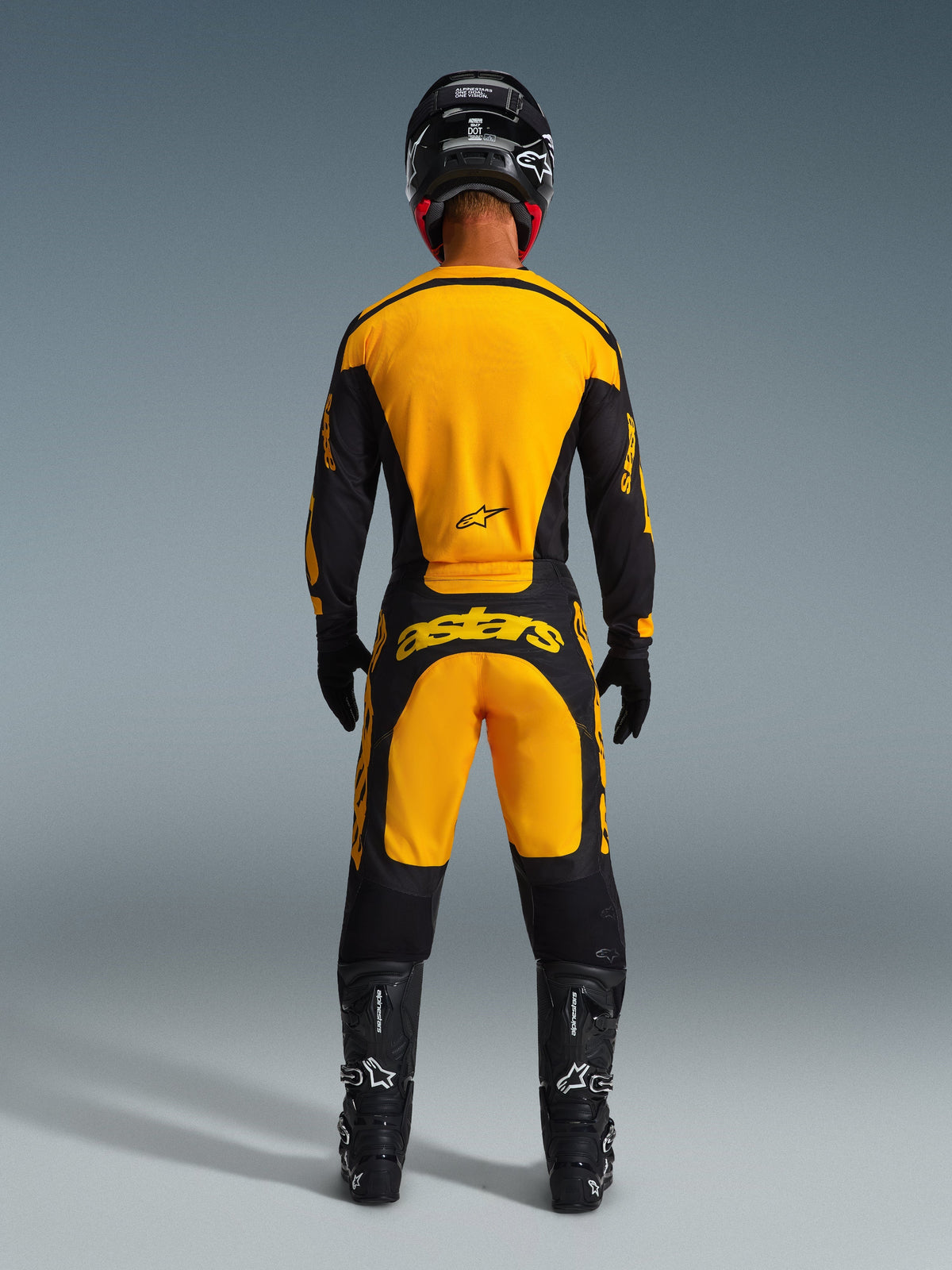 Pilote de motocross de dos portant le maillot Racer Riway 2026 Alpinestars et le pantalon assorti jaune et noir, casque noir, bottes et gants noirs, tenue de course tout-terrain complète avec logos Alpinestars.
