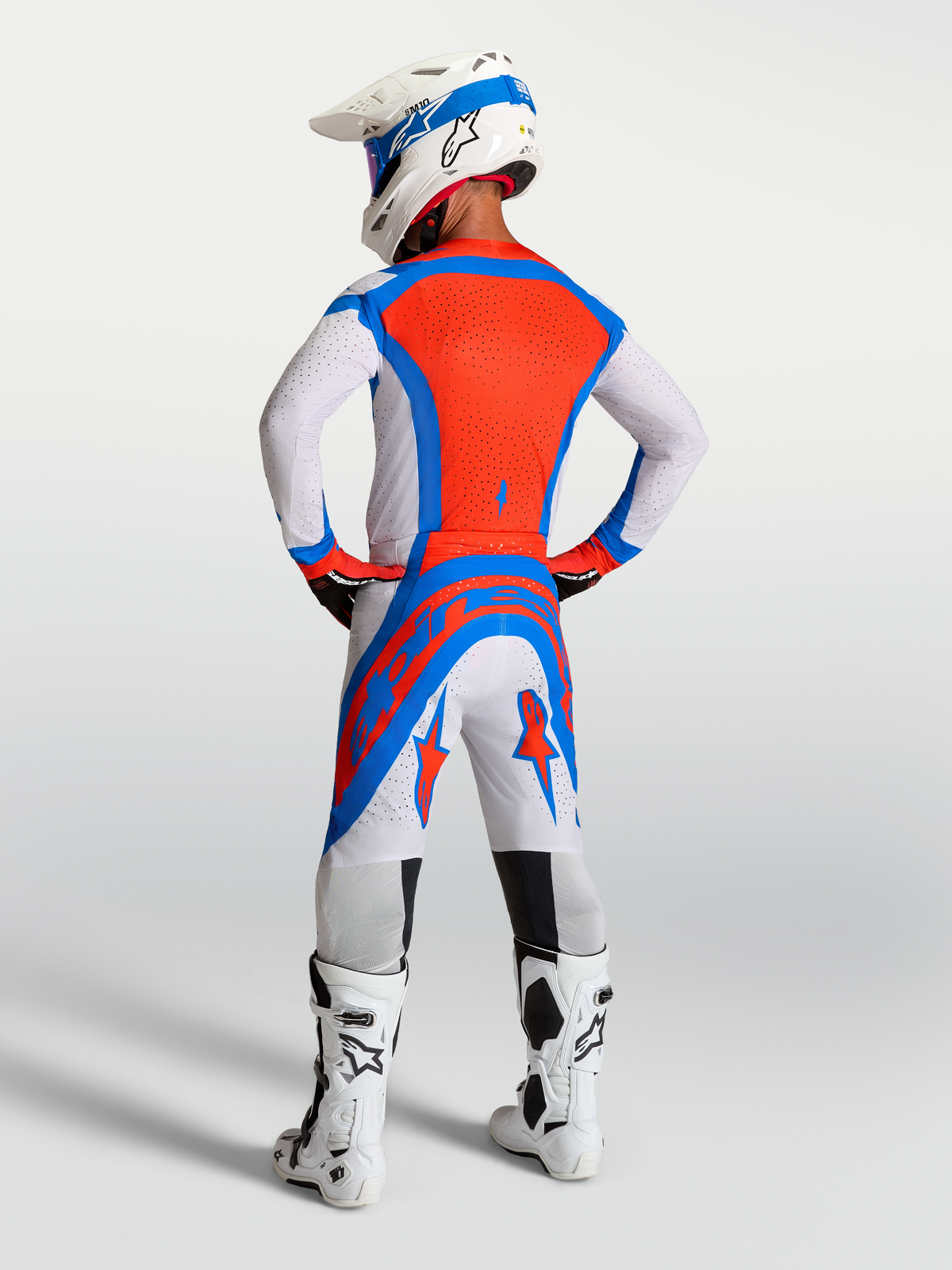 Ensemble de motocross Alpinestars Supertech Vesta, lilas clair et rouge cardinal, vue arrière d'un pilote avec casque et bottes, équipement de protection tout-terrain.