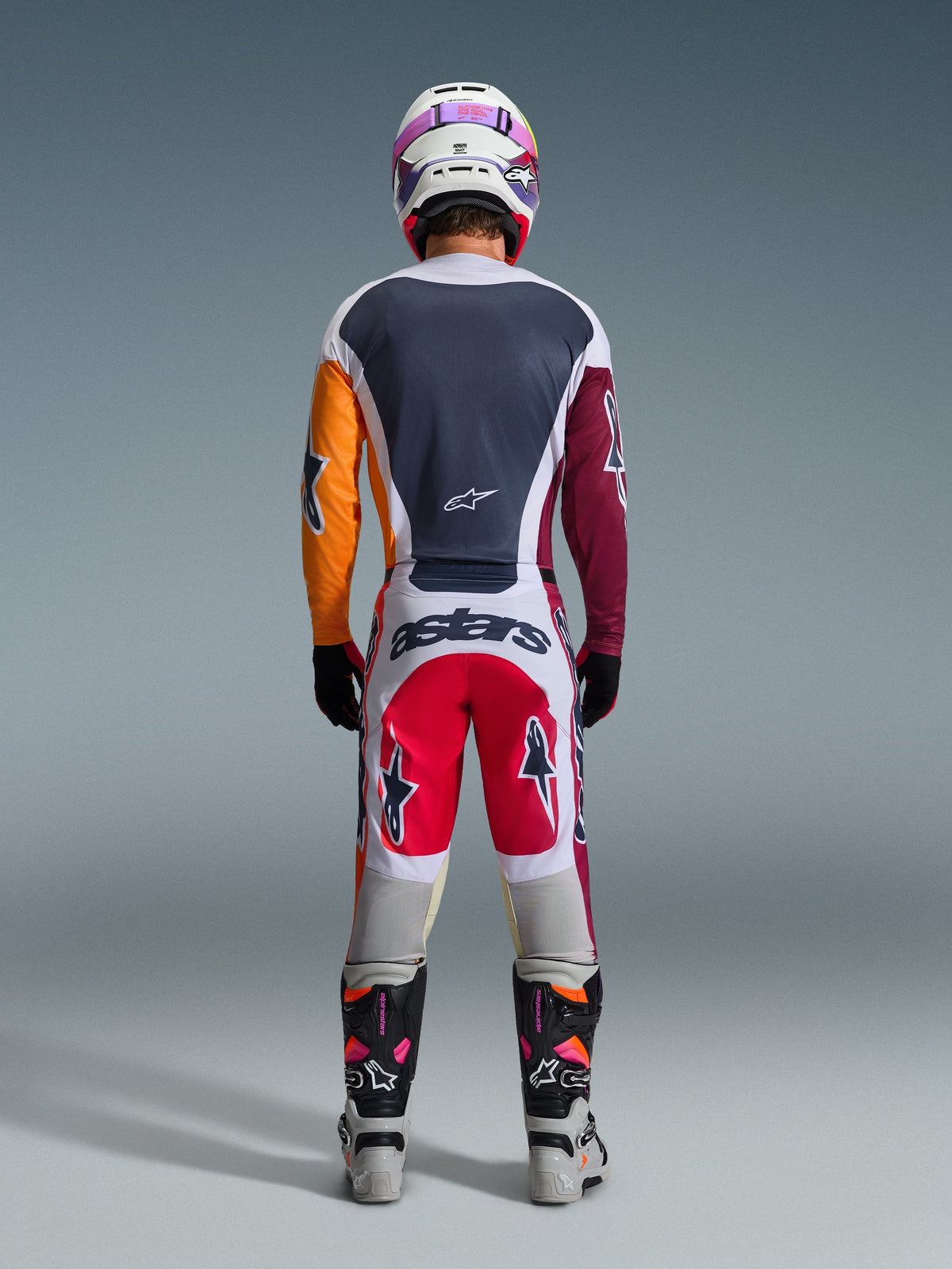 Back view of a motocross rider wearing the Alpinestars 2026 Racer Portl Gear Set, multi-colored jersey with orange and maroon sleeves, white and red pants with 'astars' branding on the seat, coordinated white helmet and technical racing boots, designed for off-road motocross performance.