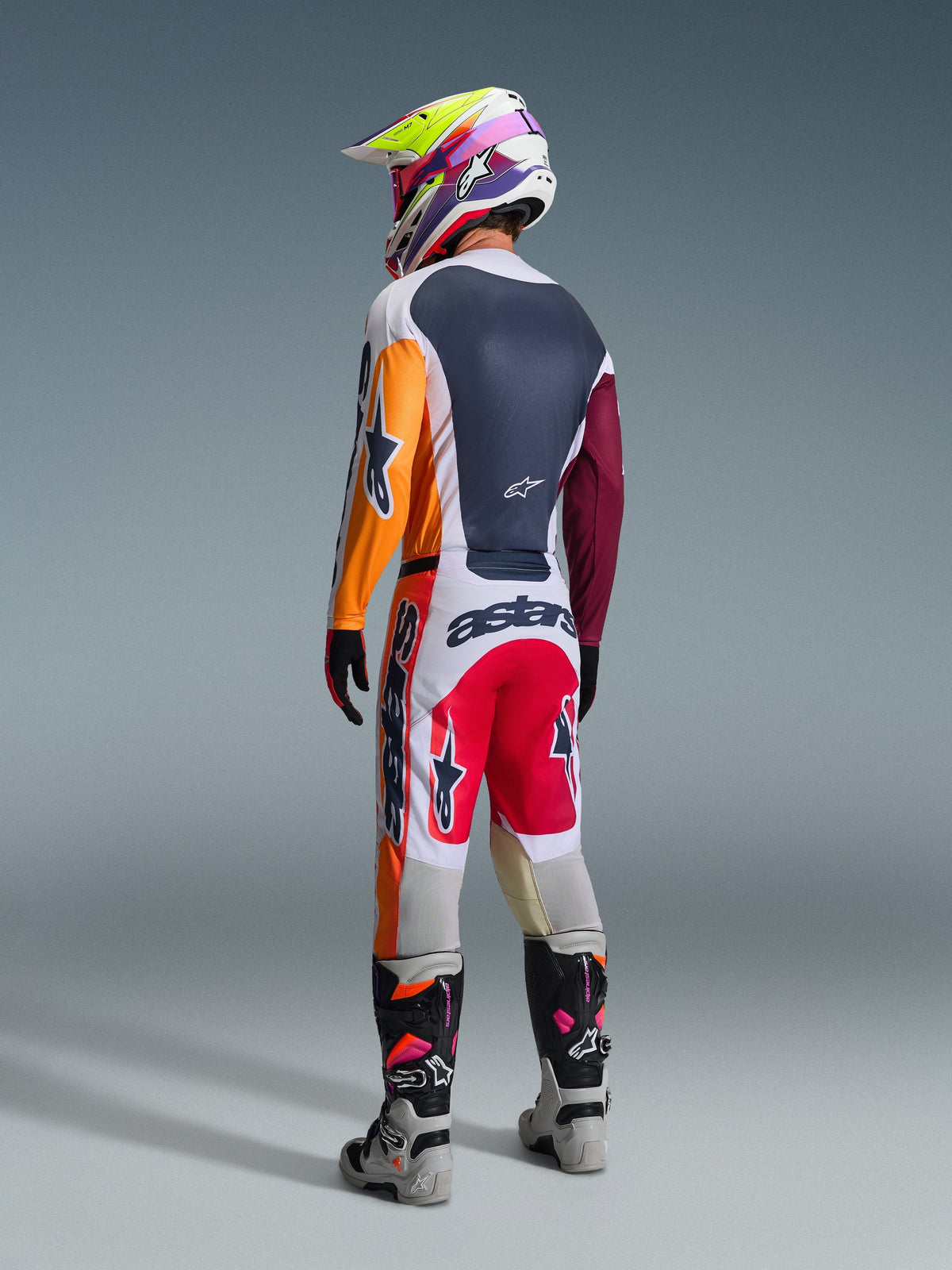 Pilote de motocross portant une tenue de course complète vue de dos, maillot Alpinestars 2026 Racer Portl en rouge, gris clair et vert foncé avec empiècements orange et bordeaux, pantalon assorti blanc et rouge, bottes grises et noires avec détails roses, casque multicolore, tenue de course tout-terrain coordonnée.