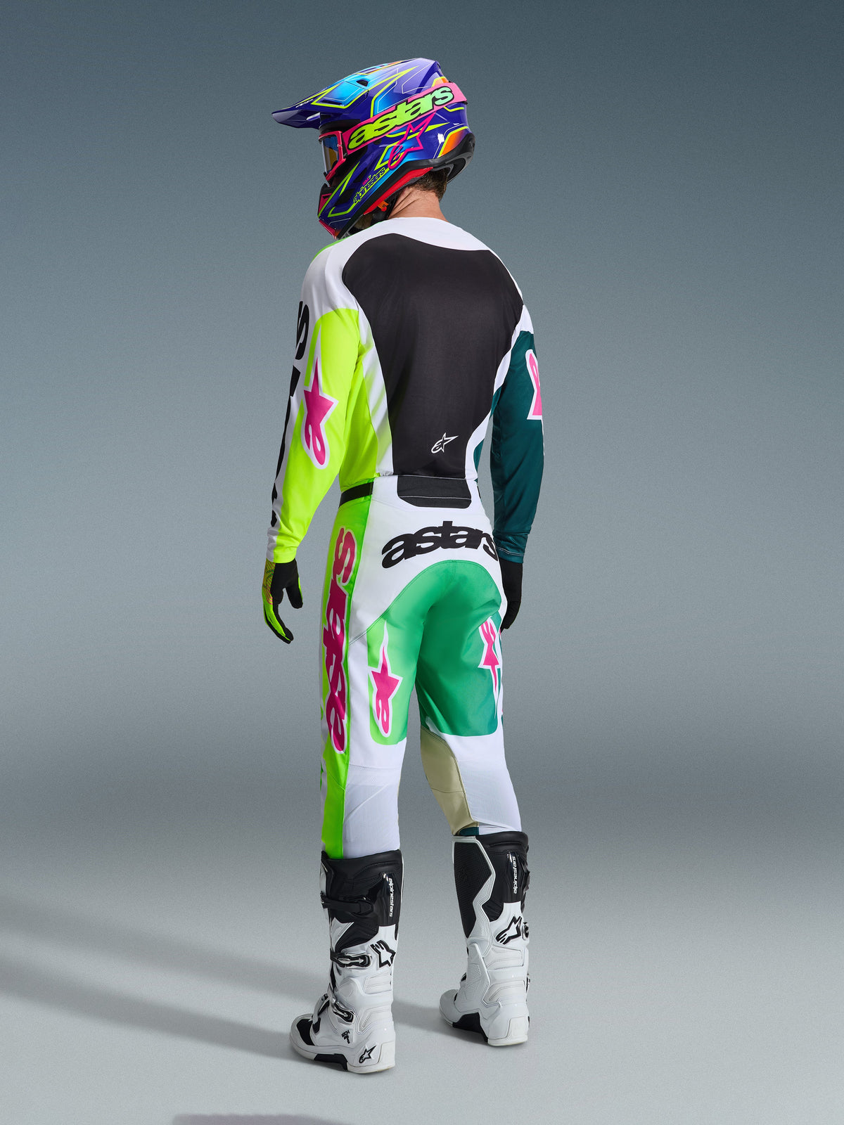 Pilote de motocross vu de dos portant la tenue Alpinestars 2026 Racer Portl, maillot avec manches jaune fluo et bleu canard et logos roses, pantalon assorti blanc et vert avec accents roses vifs, casque coloré et bottes blanches, équipement de course tout-terrain professionnel
