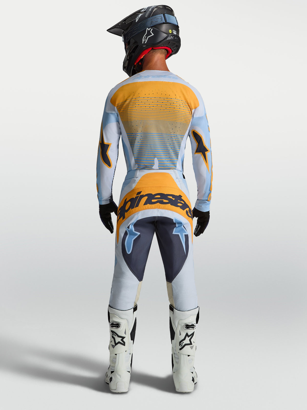 Alpinestars Maillot Techstar Maven et Techstar Maven Gear Set, équipement de motocross pour homme, bleu et orange, vue de dos, porté par un pilote avec casque et bottes de moto blanches.
