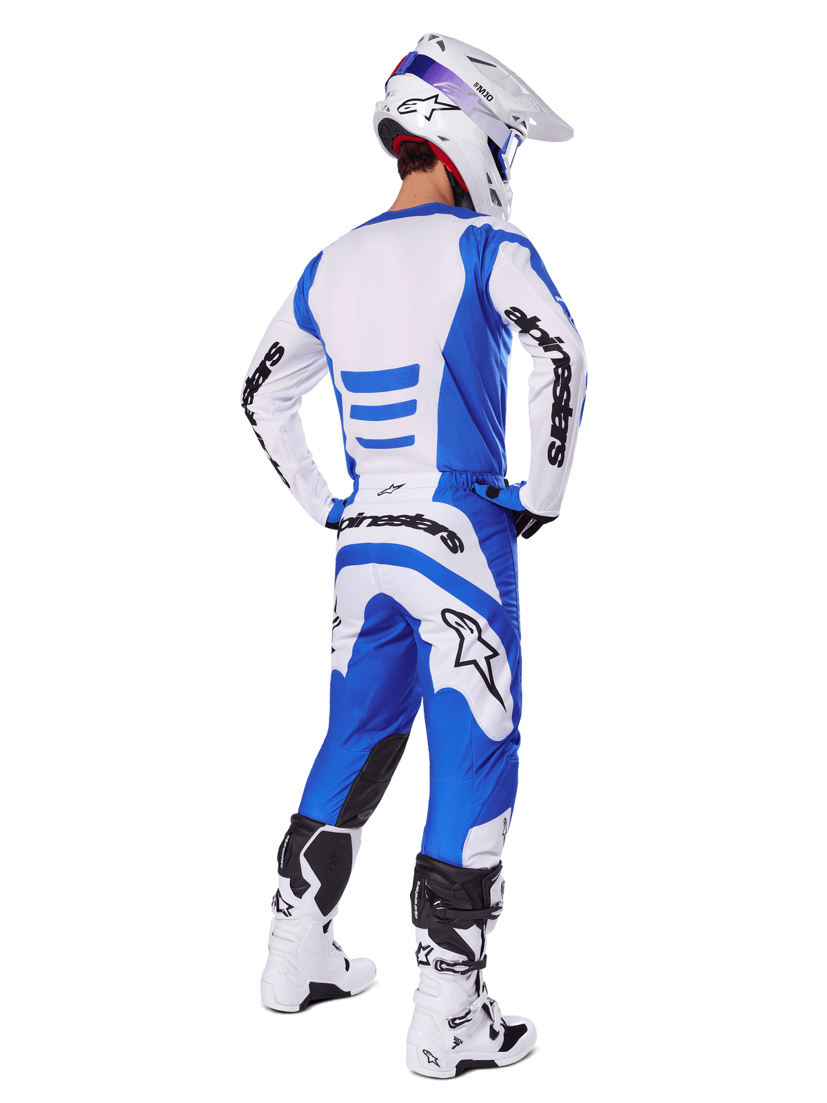Pilote de motocross de dos portant l'ensemble d'équipement Alpinestars Fluid Haul 2025, maillot bleu et blanc avec pantalon assorti, casque blanc avec accents bleus et bottes de course blanches et noires, tenue de course tout-terrain coordonnée