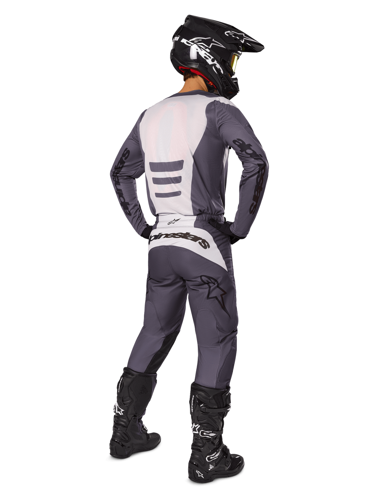 Pilote de motocross de dos portant l'ensemble d'équipement Alpinestars Fluid Haul 2025, maillot gris et blanc avec pantalon assorti, casque noir avec lunettes jaunes, bottes de motocross noires, équipement de course complet pour la performance tout-terrain