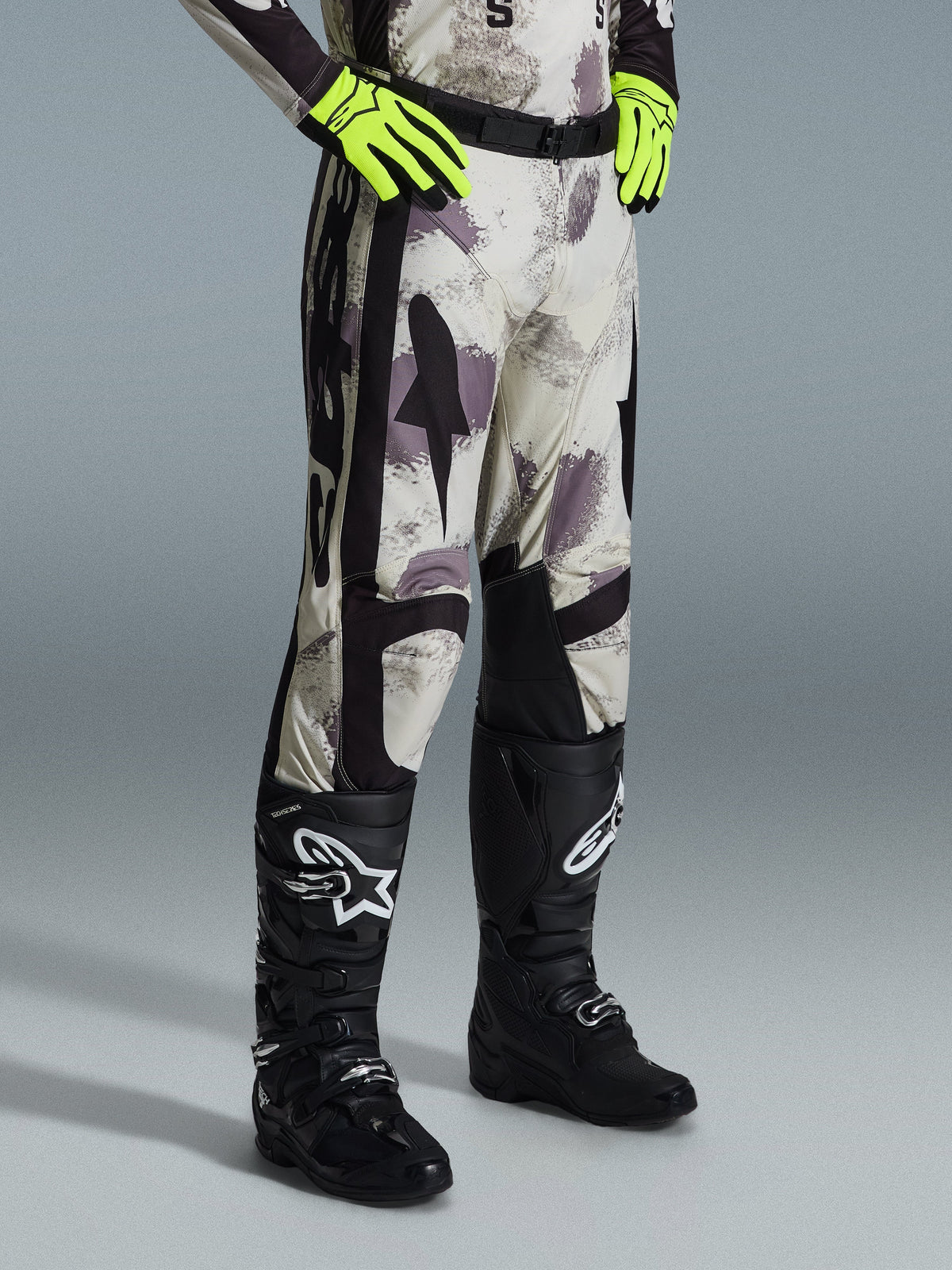 Alpinestars 2026 Racer Lahnd Pants, pantalon de motocross, Sand Camo, porté avec des gants jaune fluo et des bottes de motocross noires, présentant un motif camouflage abstrait avec des panneaux latéraux noirs, design tout-terrain ergonomique