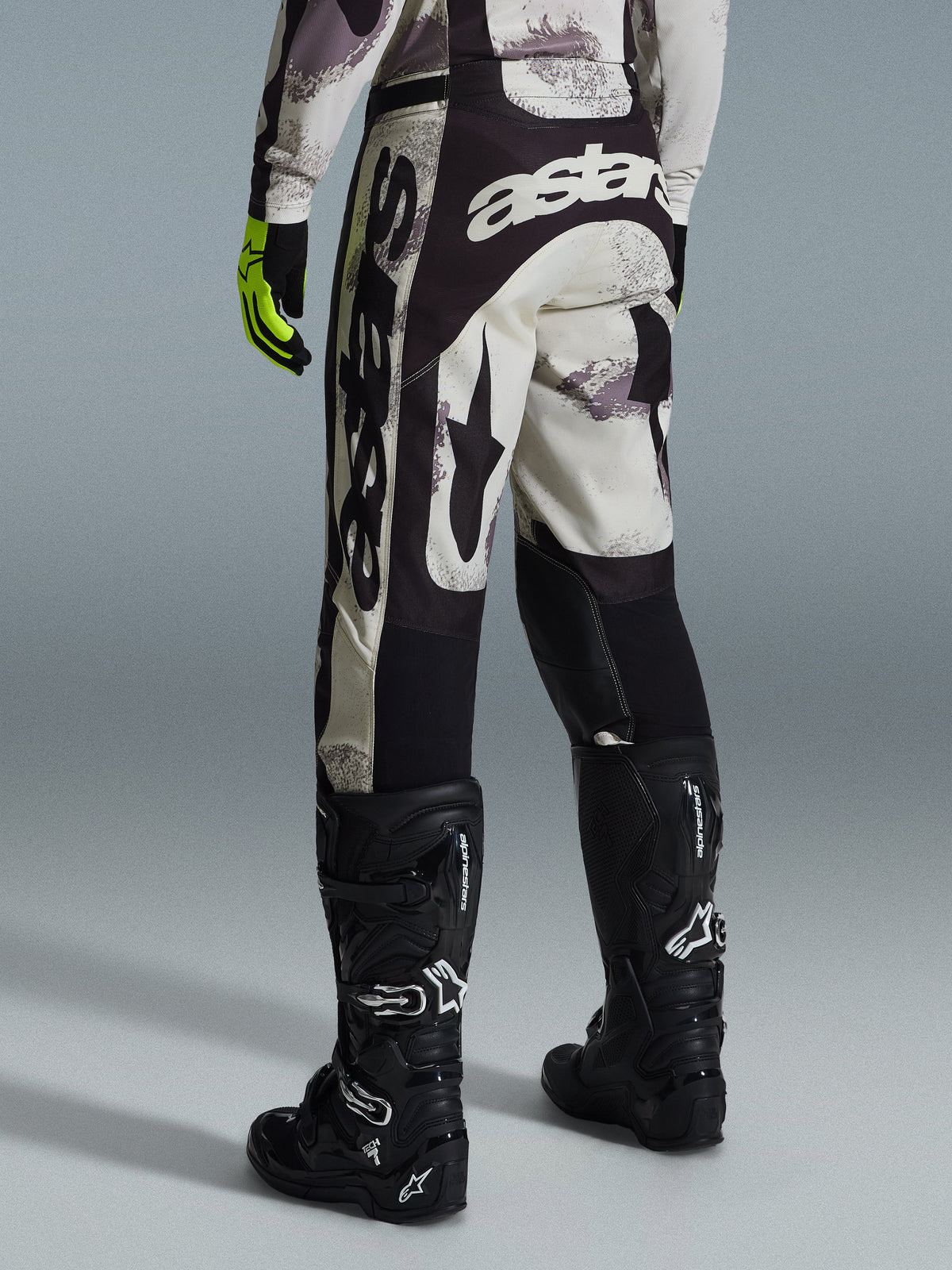 Alpinestars 2026 Racer Lahnd Pants, pantalon de motocross, Sand Camo et gris, vue arrière avec logo blanc imposant sur l'assise et les jambes, associé à des bottes de motocross noires et des gants jaunes, équipement de course tout-terrain professionnel