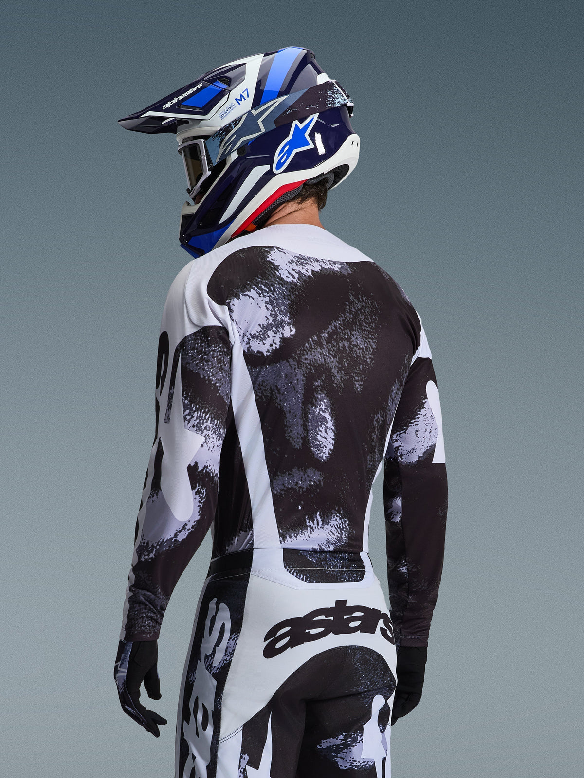 Maillot Alpinestars Racer Lahnd 2026 en Iron Camo, vue arrière d'un pilote de motocross portant une tenue à motif abstrait noir, blanc et gris, incluant le pantalon assorti, des gants noirs et un casque de course bleu avec lunettes.