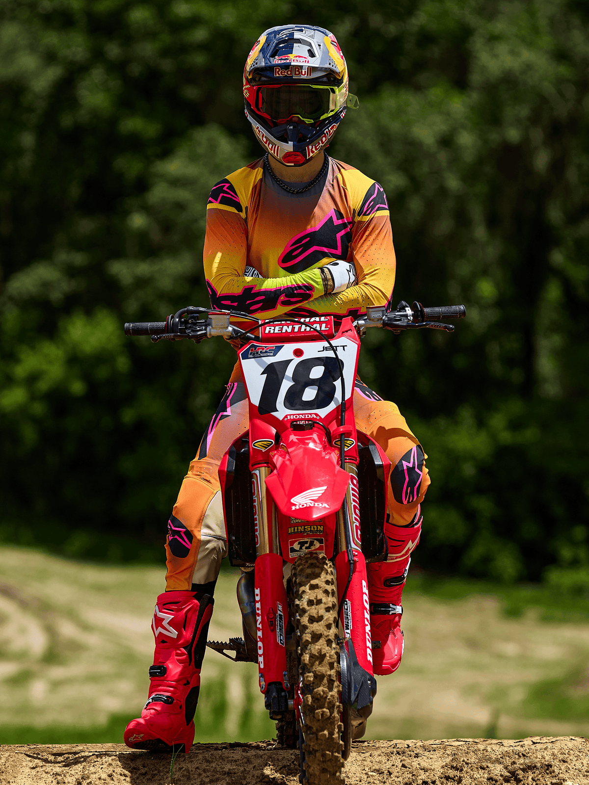 Pilote de motocross assis sur une moto Honda rouge portant le numéro 18, vêtu du maillot Alpinestars 2025 Supertech Lipan et du pantalon assorti en jaune et rose, bottes Alpinestars rose vif, casque Red Bull, équipement de course professionnel pour la performance tout-terrain.