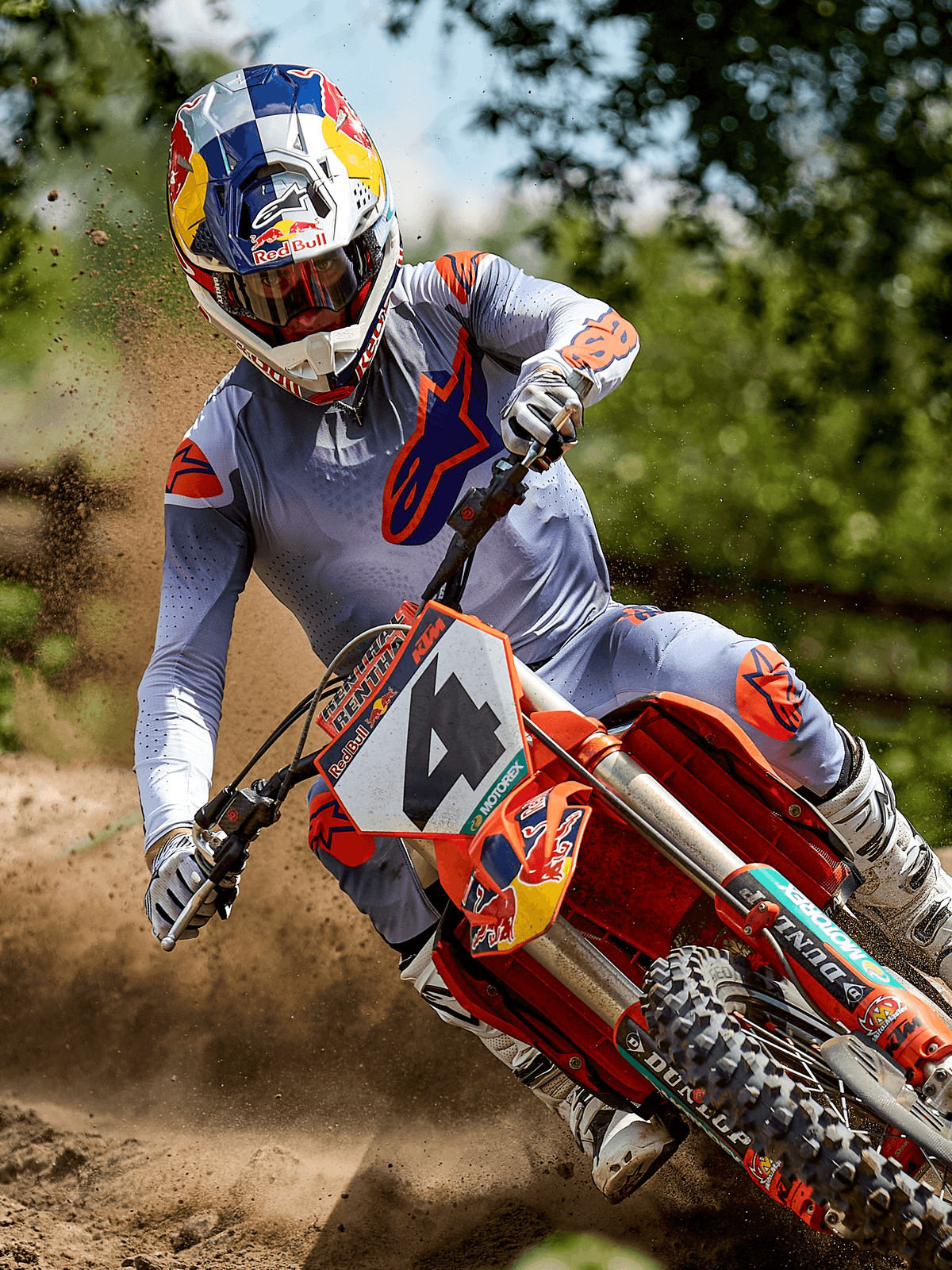 Motocross rider in action wearing the Alpinestars 2025 Supertech Lipan Gear Set in Gray Orange Fluo, featuring a light gray jersey and pants with vibrant orange Astars logos, white boots, and a Red Bull branded helmet while riding an orange KTM bike on a dirt track.
