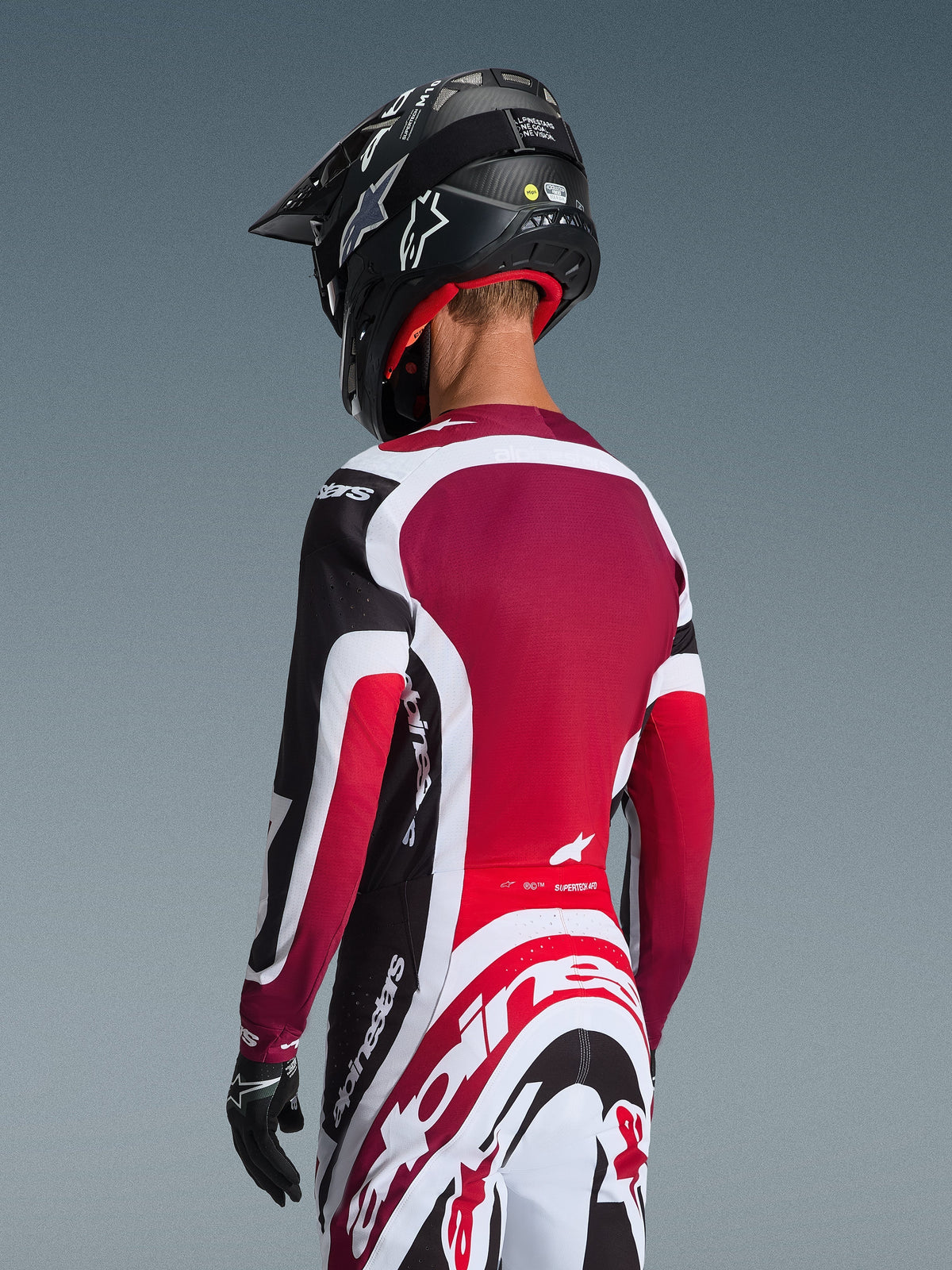 Ensemble d'équipement Alpinestars Supertech AFD Vista 2026, vue arrière d'un pilote de motocross en tenue de course noire, rouge et blanche, maillot avec panneau arrière rouge foncé et coupe athlétique, pantalon assorti avec logo Alpinestars imposant, casque et gants noirs, tenue de course de motocross professionnelle