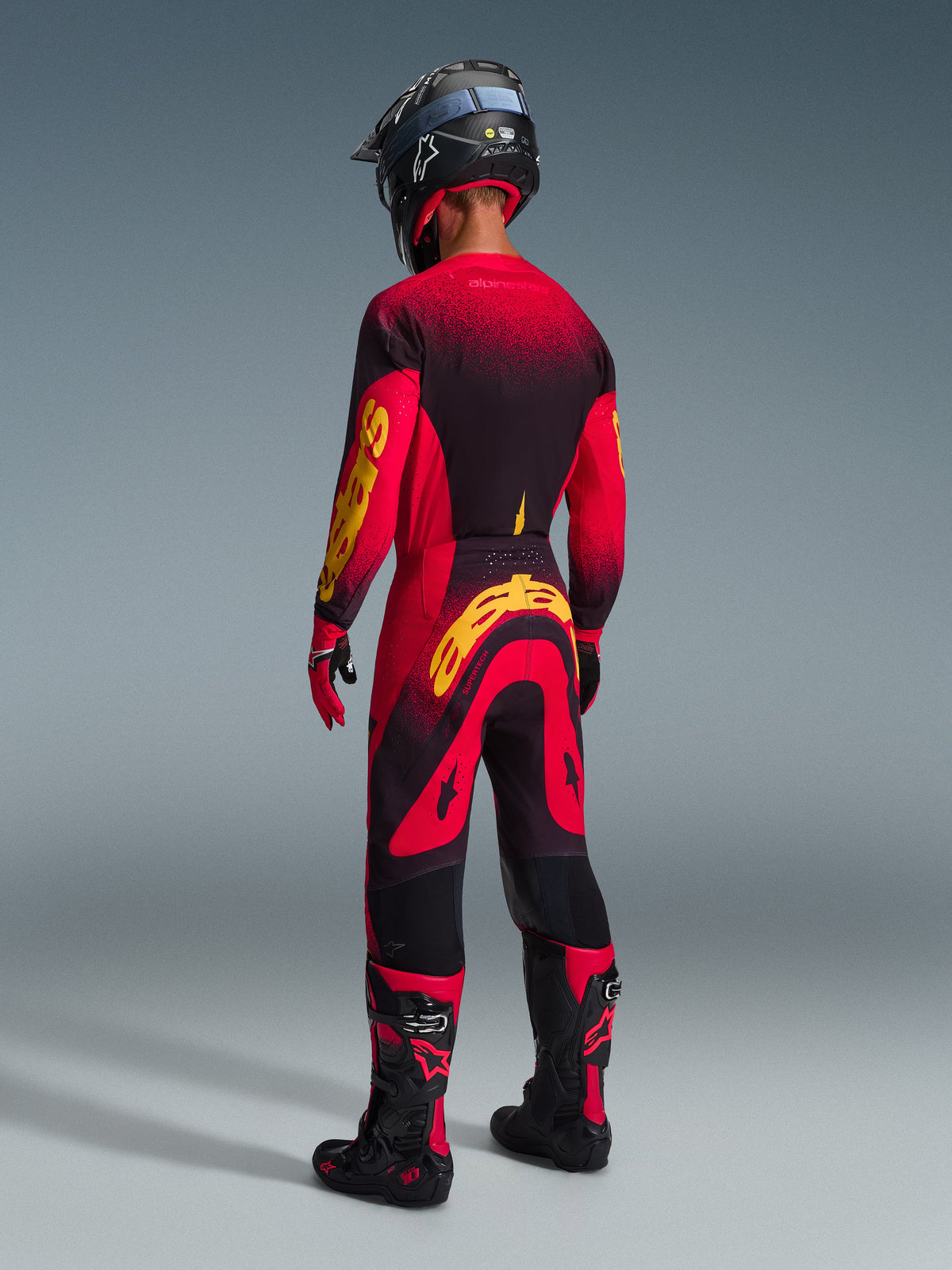 Alpinestars 2026 Supertech Scenz Gear Set, motocross rider viewed from behind, red and black jersey with yellow branding, matching red and black pants with yellow logo, red gloves, black and red motocross boots, and black helmet, coordinated pro-level racing gear.