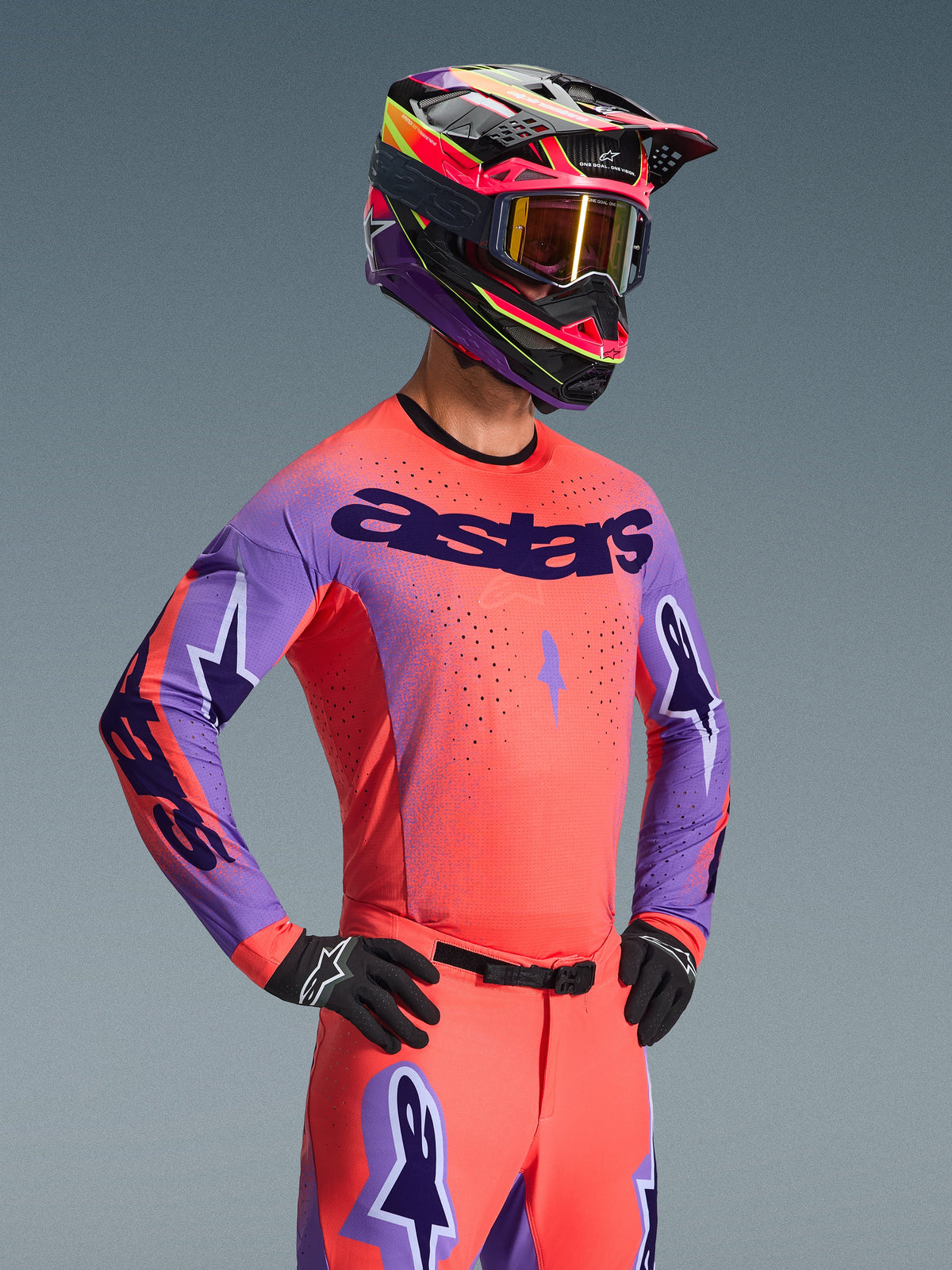 Pilote de motocross portant le maillot Alpinestars Supertech Scenz 2026, Hot Coral et violet avec un marquage 'astars' audacieux sur la poitrine, tenue assortie comprenant un pantalon corail et un casque coordonné violet et corail, présentant des perforations au laser pour la course tout-terrain haute performance.
