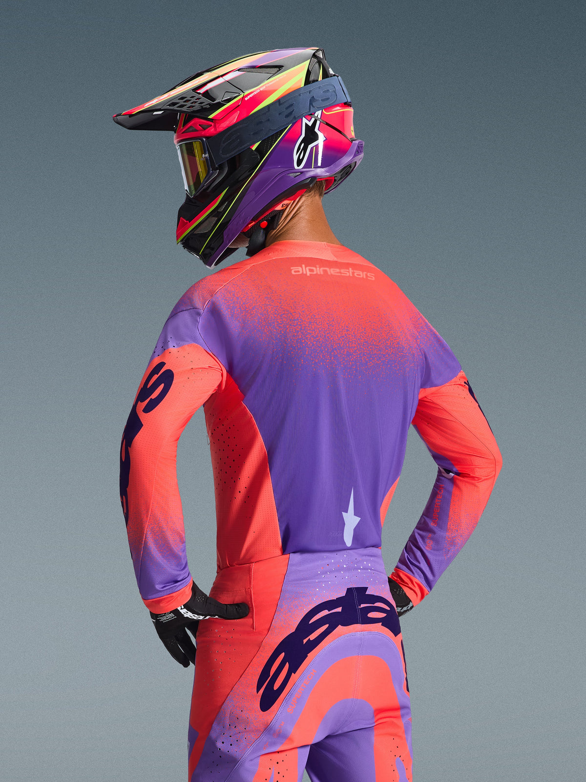 Pilote de motocross vu de dos portant le maillot Alpinestars Supertech Scenz 2026 en Hot Coral et Violet, avec un design dégradé moucheté dynamique, accompagné d'un pantalon de motocross assorti et d'un casque de course coloré, équipement professionnel pour la performance tout-terrain.