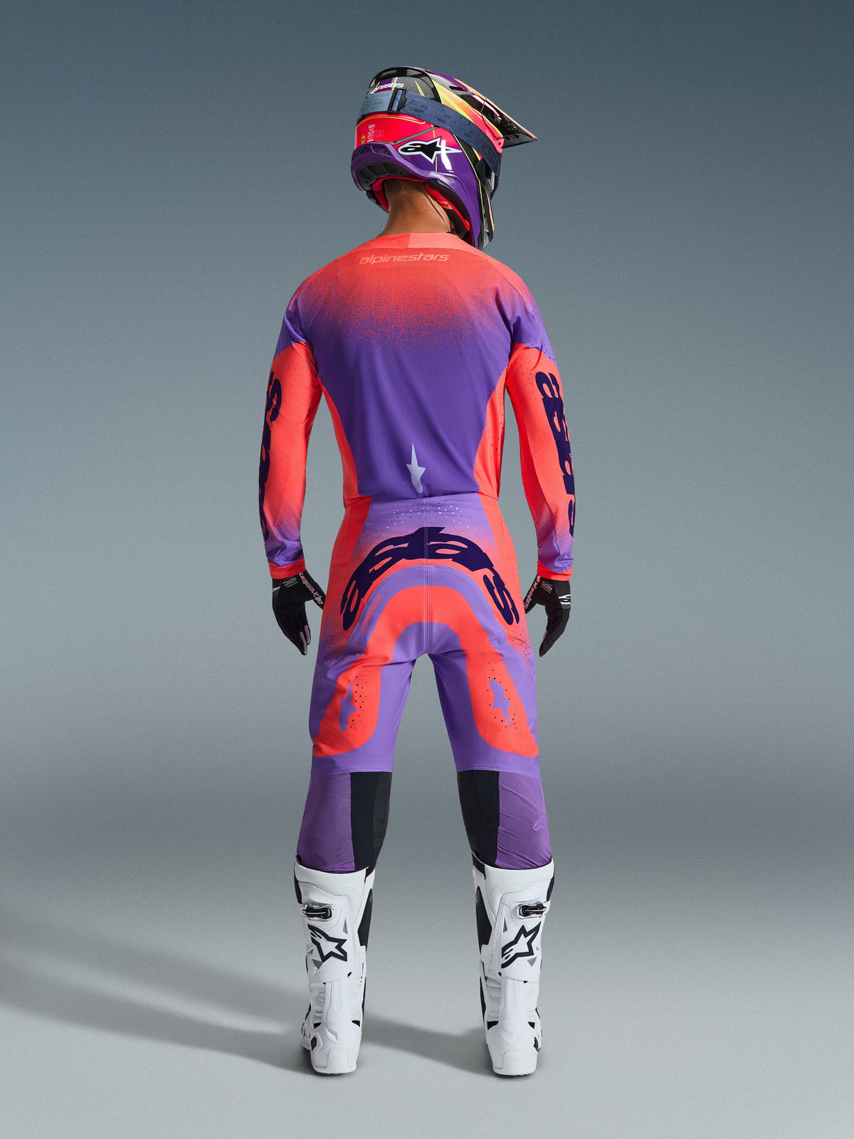 Pilote de motocross vu de dos portant l'ensemble Supertech Scenz 2026 en Hot Coral Purple, maillot et pantalon éclatants avec dégradé, logos Alpinestars, casque assorti, bottes blanches et noires et gants noirs, équipement professionnel pour le tout-terrain.
