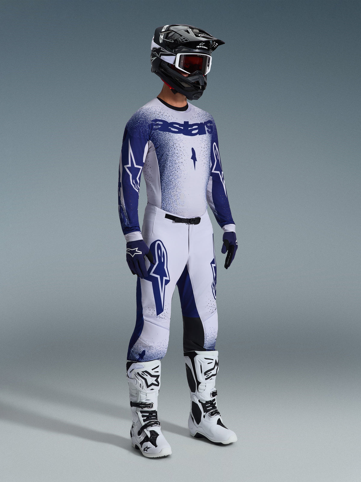 Ensemble d'équipement Alpinestars Supertech Scenz 2026, pilote de motocross portant un maillot et un pantalon de course blanc et bleu marine avec un effet de dégradé pointillé, logos Astars imposants, accompagné d'un casque noir et blanc, de gants bleu marine et de bottes de motocross blanches.