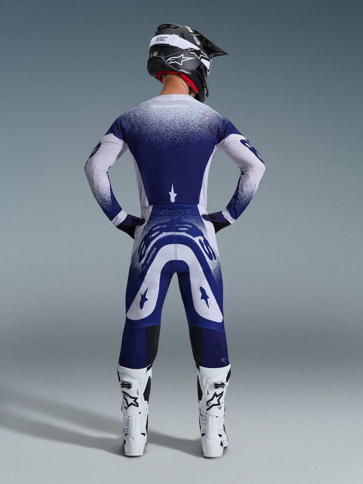 Ensemble d'équipement Alpinestars 2026 Supertech Scenz, vue arrière d'un pilote de motocross en maillot et pantalon de course bleus et blancs, effet de dégradé en pointillés, avec bottes de motocross blanches et casque noir, équipement de cross de niveau professionnel