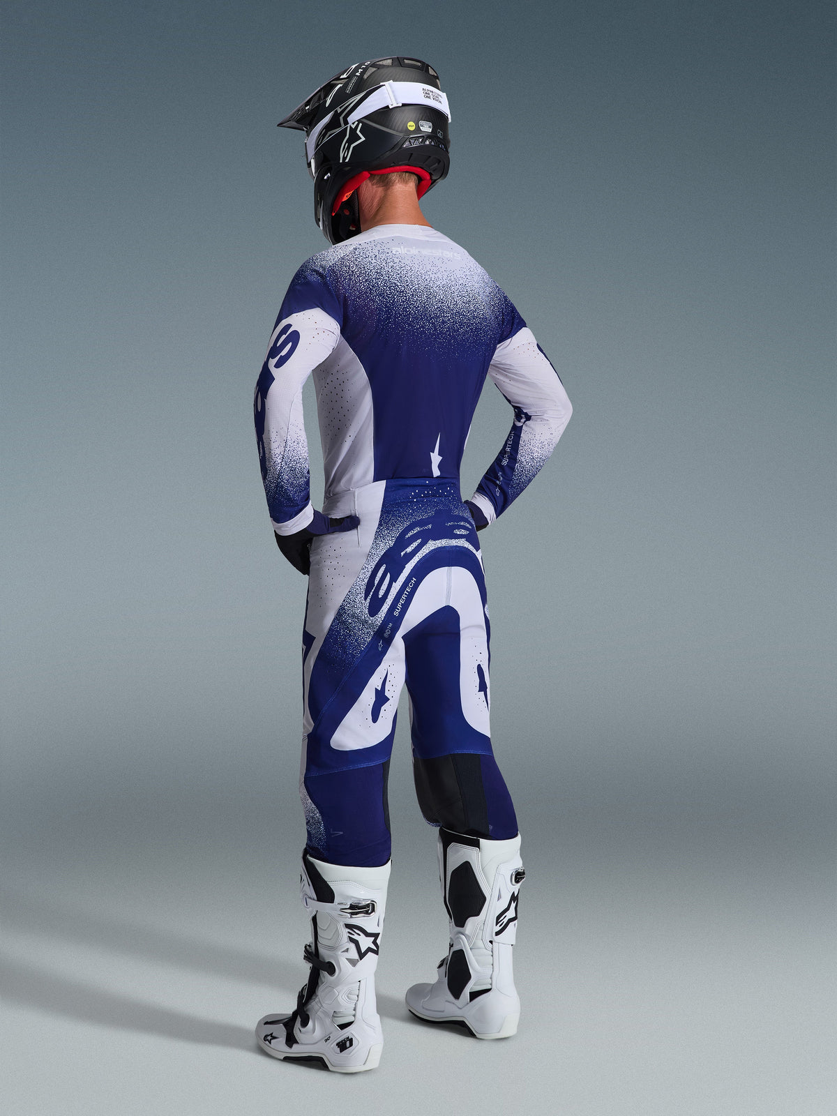 Pilote de motocross portant l'ensemble d'équipement Alpinestars 2026 Supertech Scenz, maillot et pantalon bleu marine et blanc avec un effet de dégradé moucheté, vue arrière montrant la coupe athlétique, associé à des bottes blanches et noires et un casque noir, équipement de course professionnel pour la performance tout-terrain