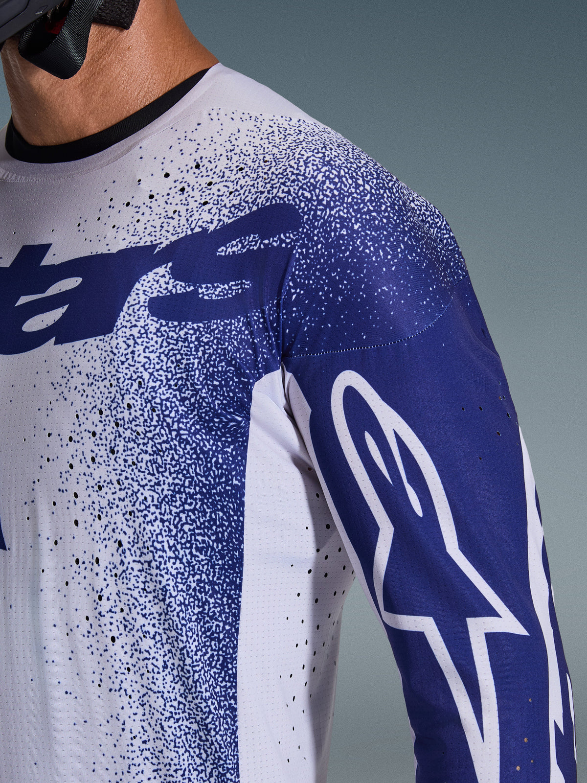 Maillot Alpinestars Supertech Scenz 2026, gris clair et bleu foncé, gros plan sur l'épaule et la poitrine montrant des perforations au laser, logo Astars blanc sur la manche bleue, design graphique moucheté, équipement de motocross professionnel.