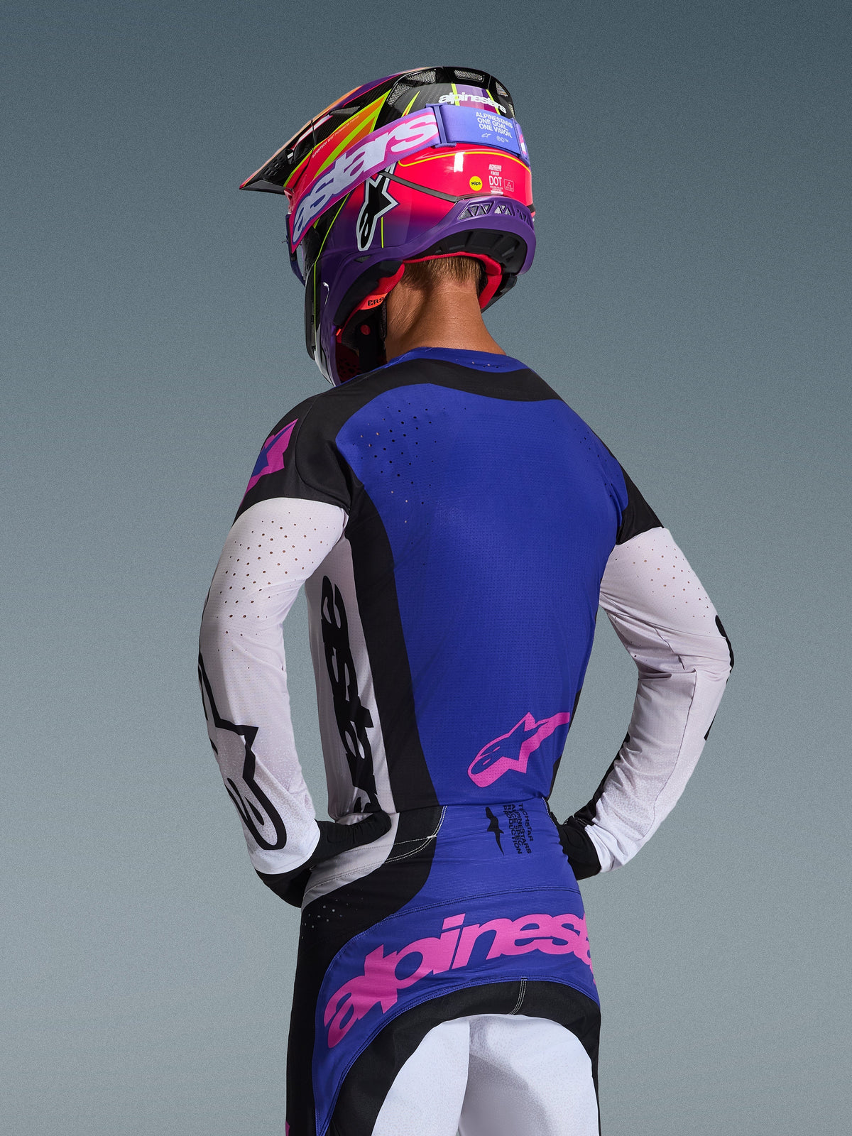 Motocross rider from the back wearing the 2026 Techstar Knif Jersey, Black Light Gray and Purple, featuring white perforated sleeves and pink Alpinestars logos, paired with a matching purple and pink helmet, professional off-road racing gear.
