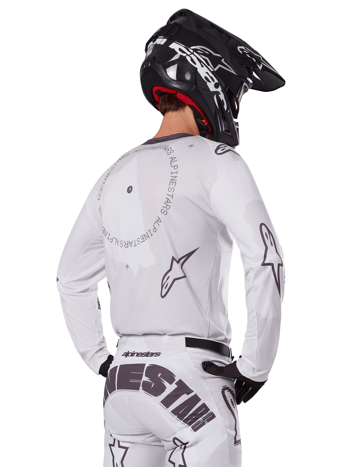 Pilote de motocross vu de dos portant l'ensemble Alpinestars 2025 Racer Hollow blanc, maillot blanc à manches longues avec logos d'étoiles gris et graphismes circulaires, pantalon blanc assorti avec marquage imposant sur l'assise, casque noir, équipement de course tout-terrain coordonné