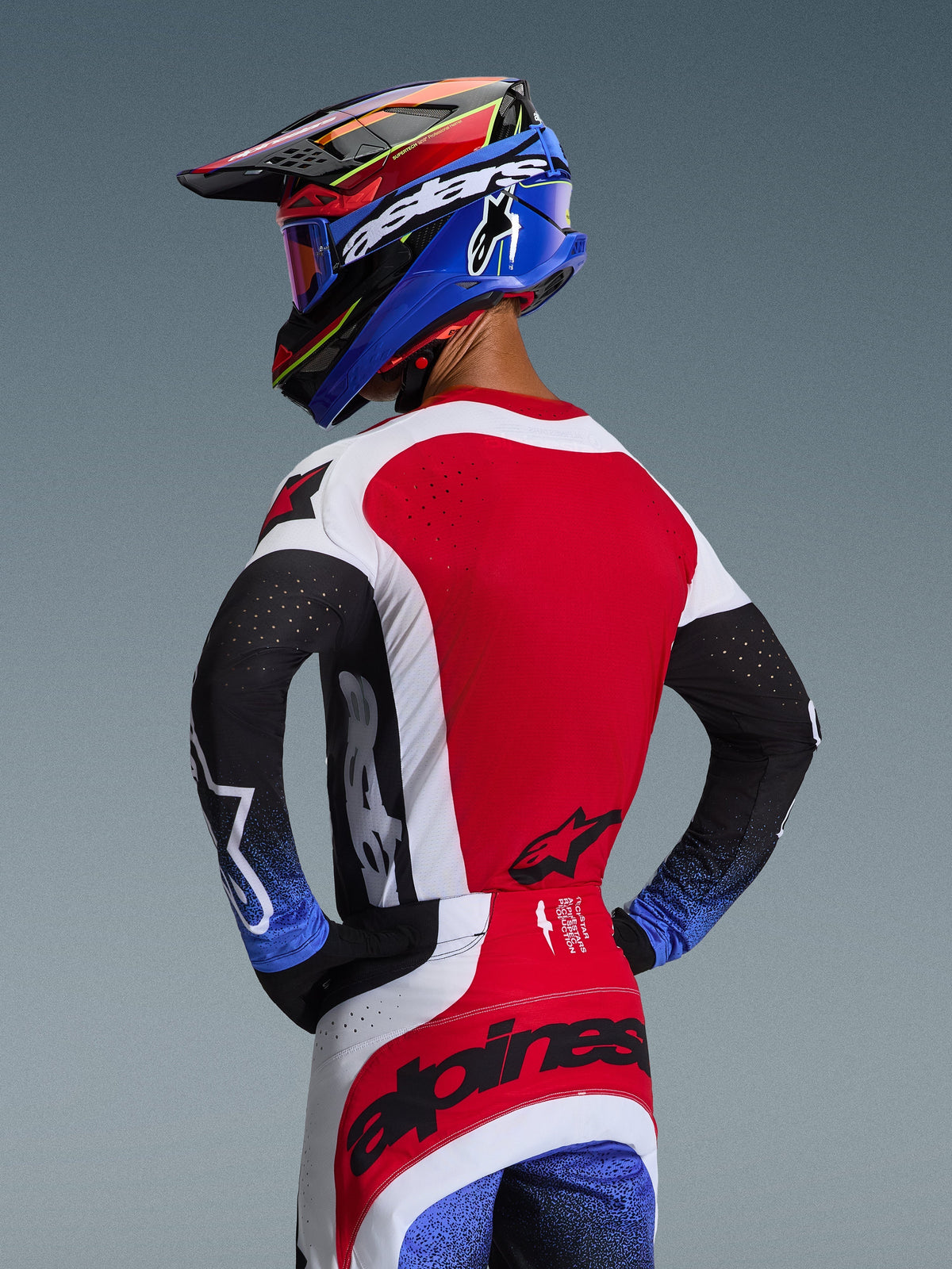 Motocross rider viewed from behind wearing the 2026 Techstar Knif Jersey in Red White and Black, featuring laser-cut perforations on the back and bold branding, paired with matching pants with blue accents and a blue and black Alpinestars helmet, professional off-road racing aesthetic