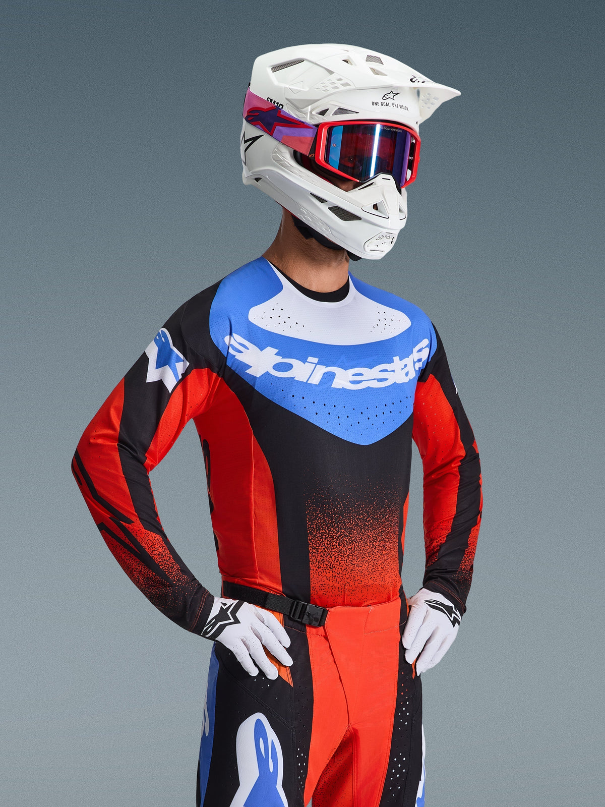 Pilote de motocross portant le maillot Techstar Knif 2026 en orange, noir et bleu UCLA, avec un empiècement bleu sur la poitrine et le logo Alpinestars blanc, des manches noires et orange avec perforations laser, accompagné d'un casque blanc, de lunettes rouges et d'un pantalon de course assorti.