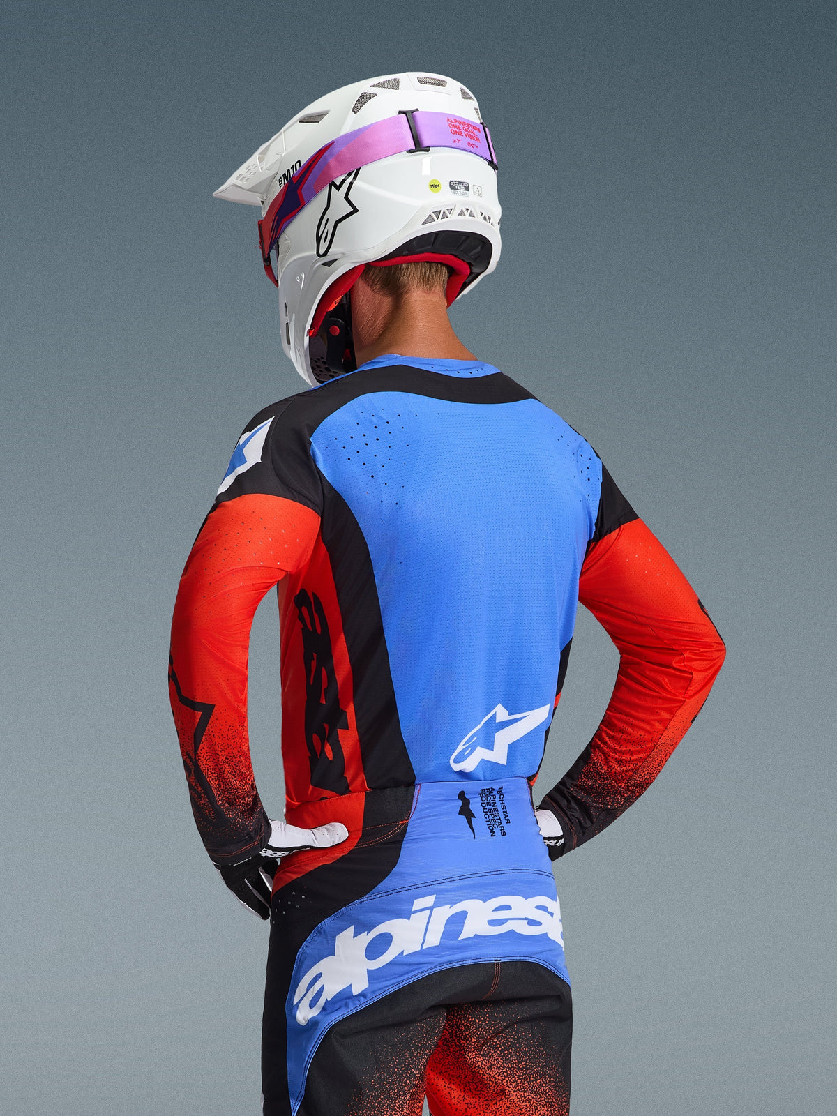 Vue arrière d'un pilote de motocross portant le maillot Techstar Knif 2026 en coloris Orange Black UCLA Blue, avec un panneau arrière bleu doté de perforations découpées au laser, des manches orange et des détails noirs, associé à un pantalon de motocross assorti et un casque Alpinestars blanc.