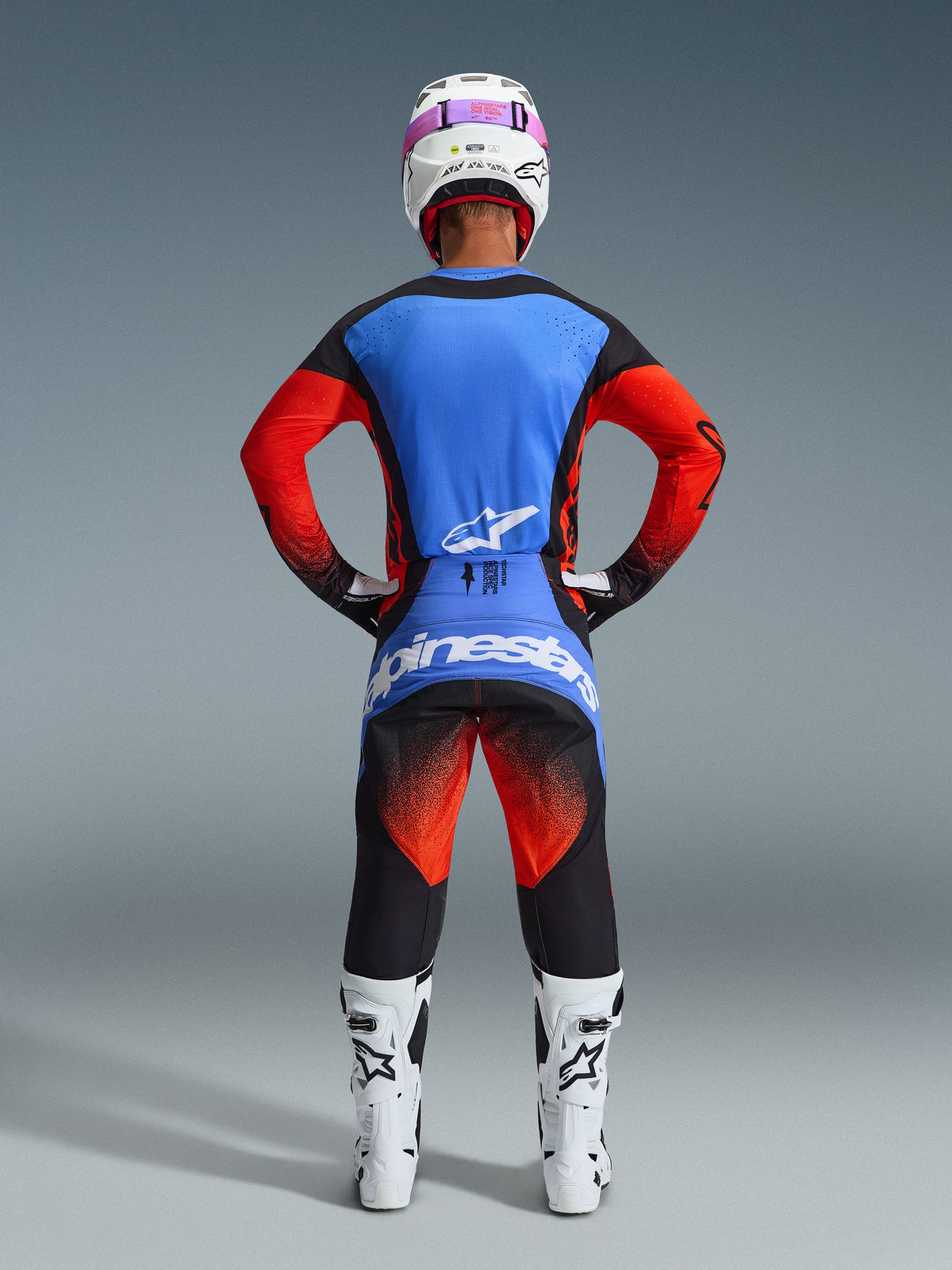 Rear view of a motocross rider wearing the Alpinestars 2026 Techstar Knif Gear Set, blue and red jersey with black accents, black pants with red gradient on the thighs and blue branding, paired with white racing boots and a white helmet with pink goggles.