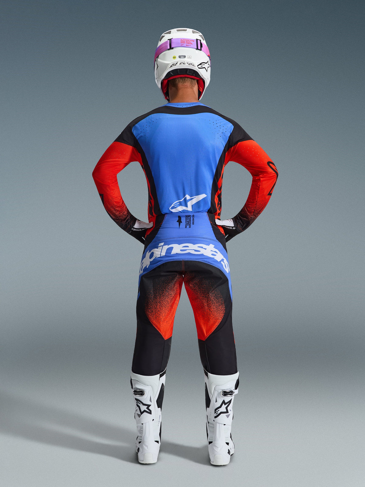 Pilote de motocross vu de dos portant une tenue de course complète, maillot Techstar Knif 2026 avec dos bleu et manches orange, pantalon noir avec accents orange et bleus, casque blanc avec sangle de lunettes rose, bottes blanches et noires, tenue coordonnée pour la course tout-terrain