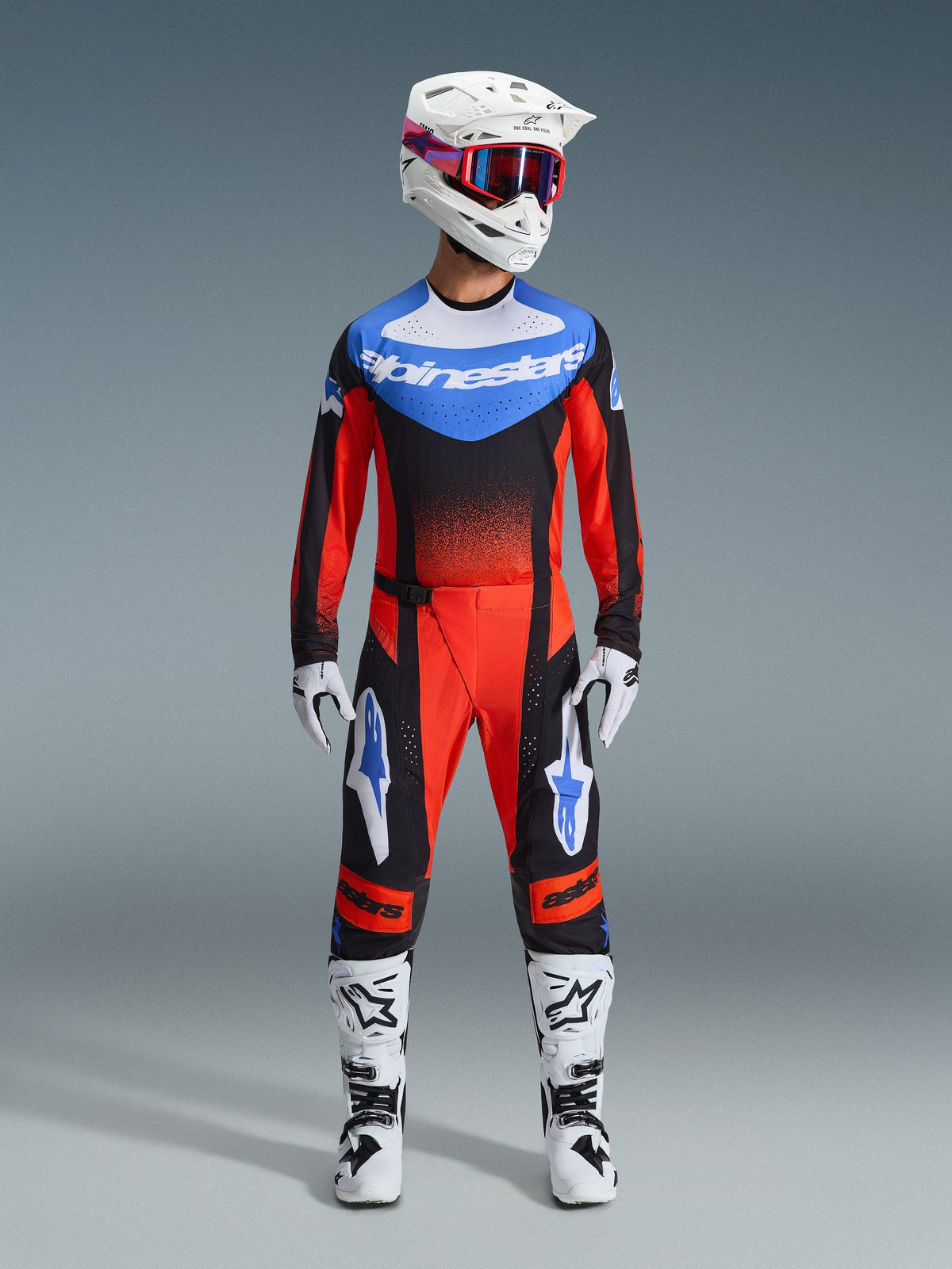 Pilote de motocross portant une tenue de course complète, maillot Techstar Knif 2026 en Orange Noir et Bleu UCLA, pantalon de motocross assorti, casque blanc avec lunettes, gants blancs et bottes blanches, tenue Alpinestars coordonnée pour la compétition tout-terrain