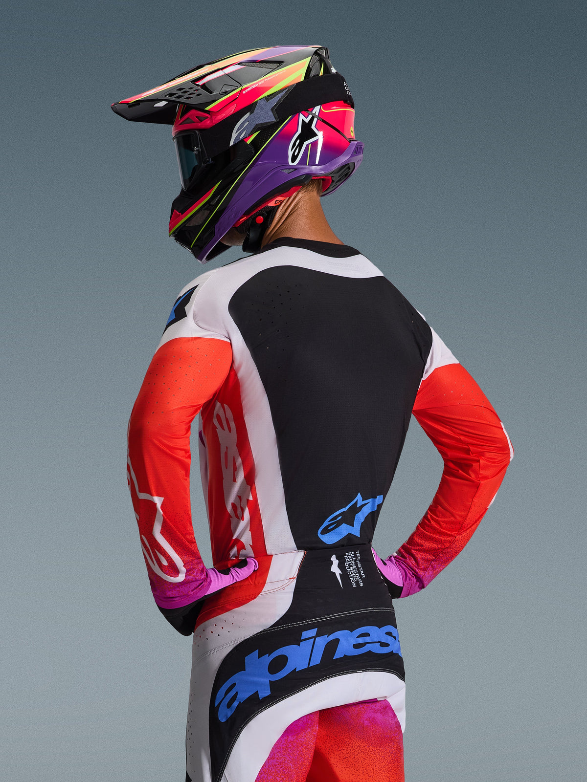Motocross rider from behind wearing Alpinestars 2026 Techstar Knif Gear Set, orange fluo sleeves with black back panel, white and black pants with blue logos, vibrant purple and pink helmet, professional motocross racing gear for off-road performance