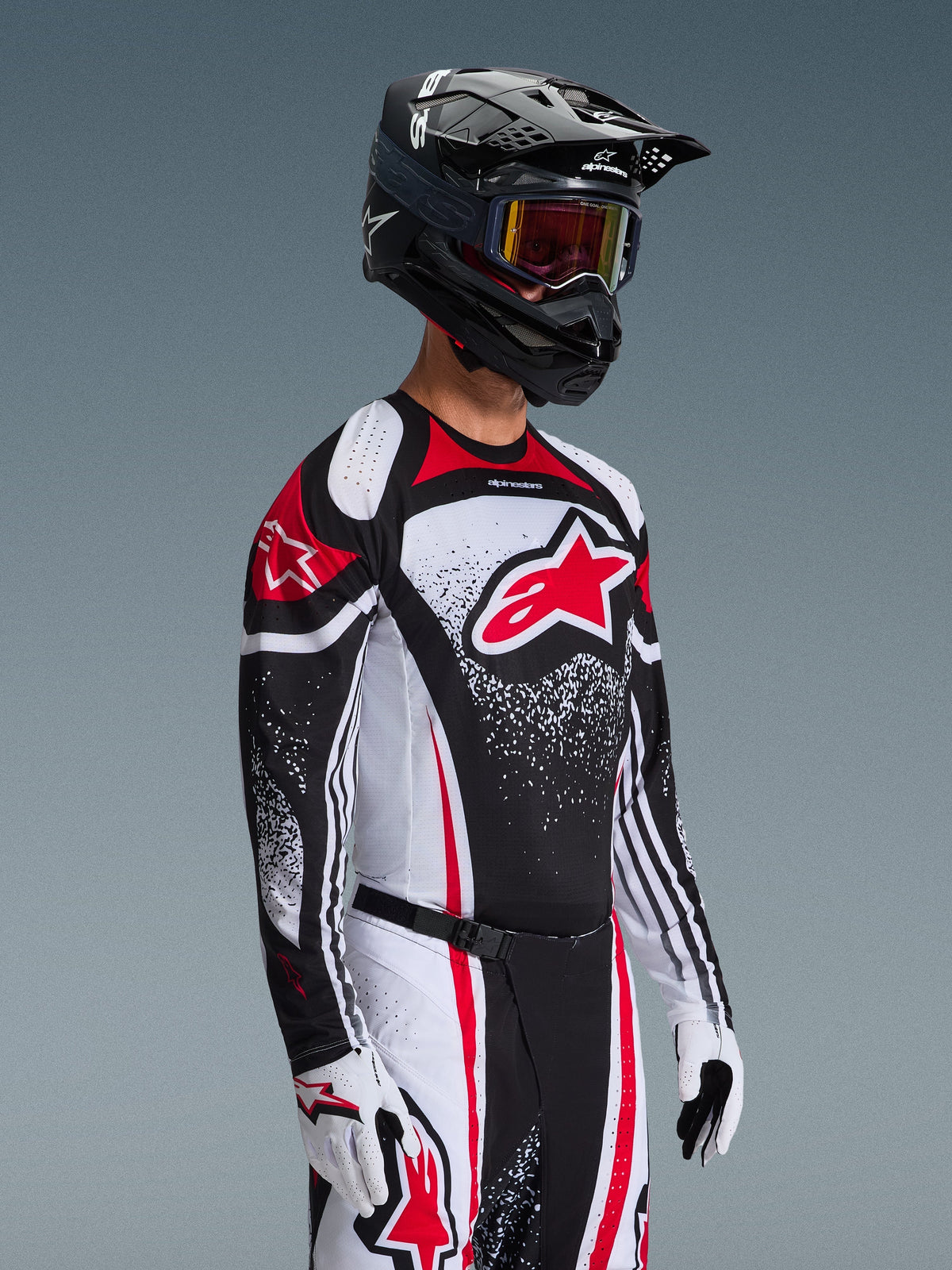 Pilote de motocross portant le maillot Alpinestars Techstar Nomur 2026 en noir, blanc et rouge, avec un grand logo Astars rouge et des motifs graphiques mouchetés, associé à un pantalon de course assorti, des gants blancs et un casque noir avec lunettes, tenue coordonnée pour la compétition tout-terrain