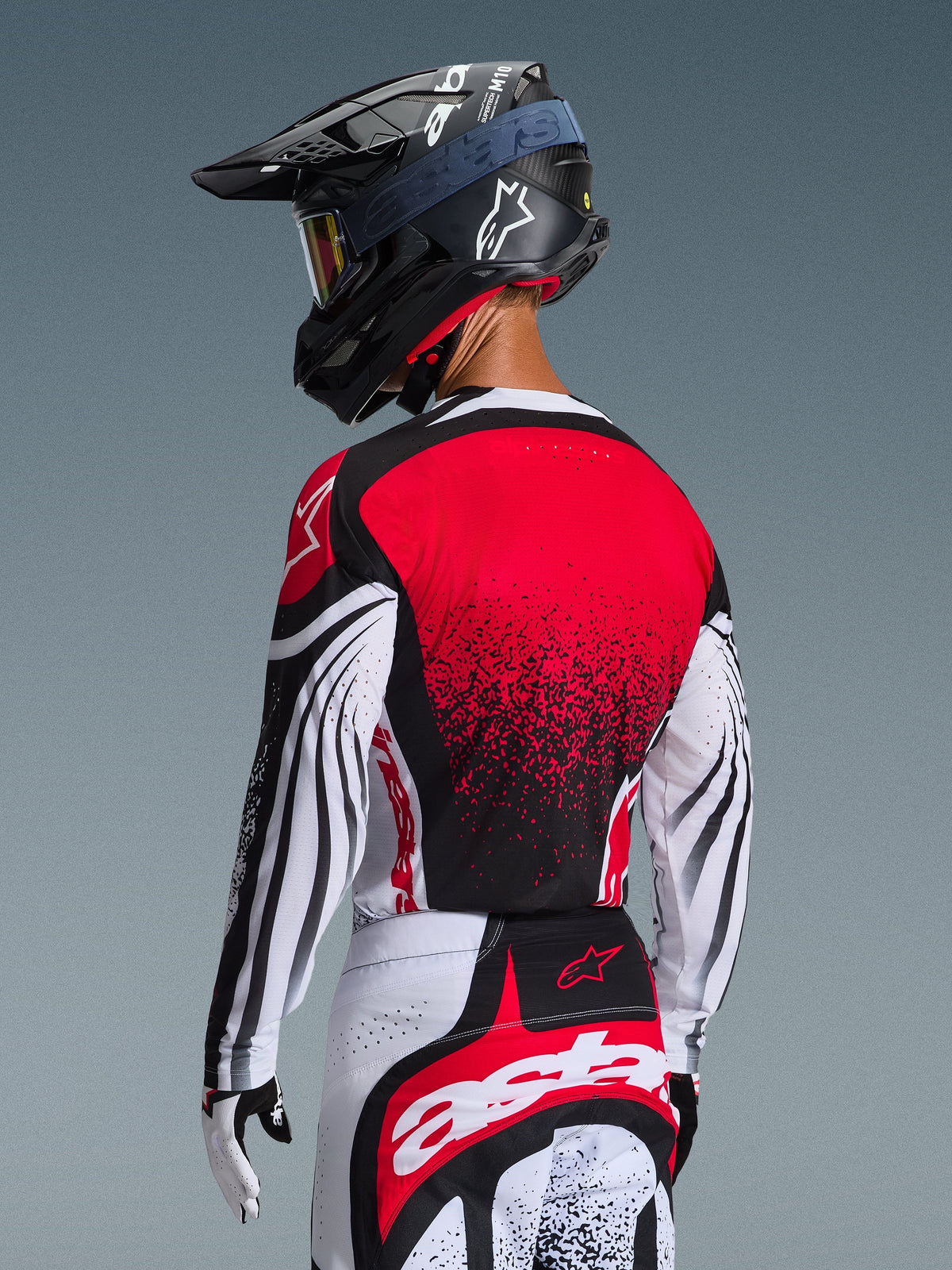 Pilote de motocross de dos portant le maillot Techstar Nomur 2026, dos rouge avec motif dégradé noir et manches blanches avec accents noirs, porté avec un pantalon rouge et blanc assorti, un casque noir et des gants blancs, équipement de course tout-terrain professionnel