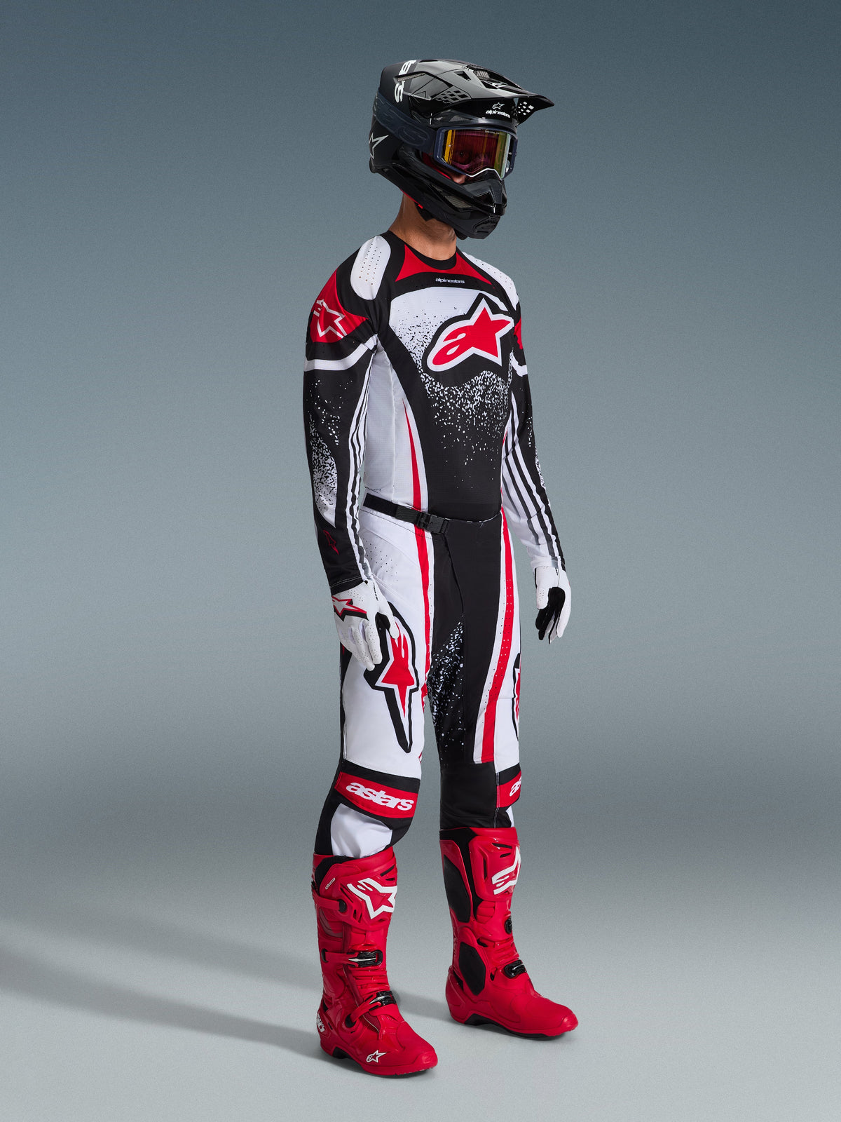 Pilote de motocross portant l'ensemble Alpinestars Techstar Nomur 2026, maillot noir avec motifs graphiques rouges et blancs, pantalon assorti, bottes rouge vif, casque noir et lunettes, tenue de course tout-terrain coordonnée