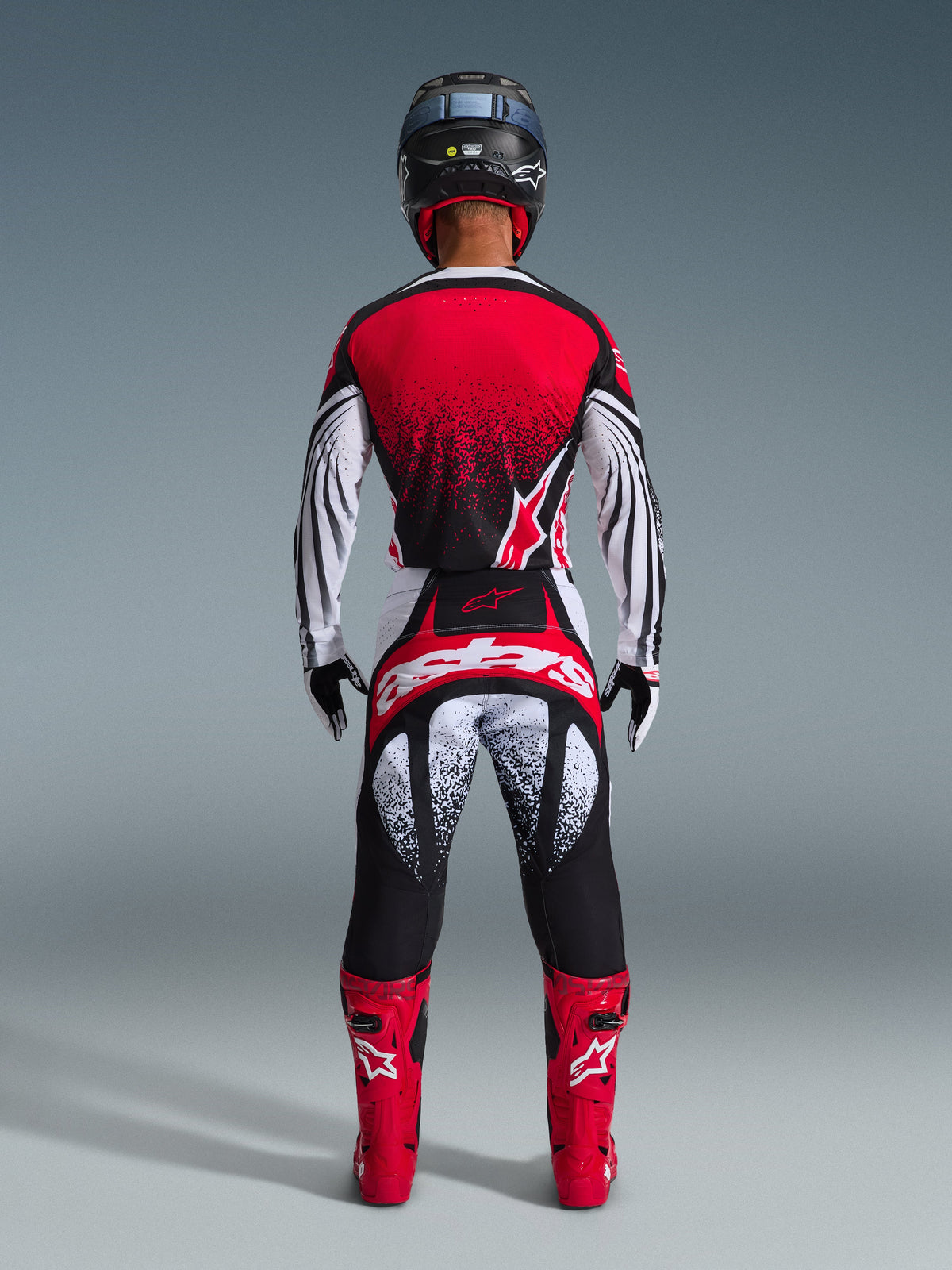 Pilote de motocross de dos portant l'ensemble Alpinestars Techstar Nomur 2026, maillot rouge et blanc avec dégradé noir moucheté, pantalon blanc et rouge assorti avec accents noirs, bottes rouge vif et casque noir, équipement de course tout-terrain coordonné
