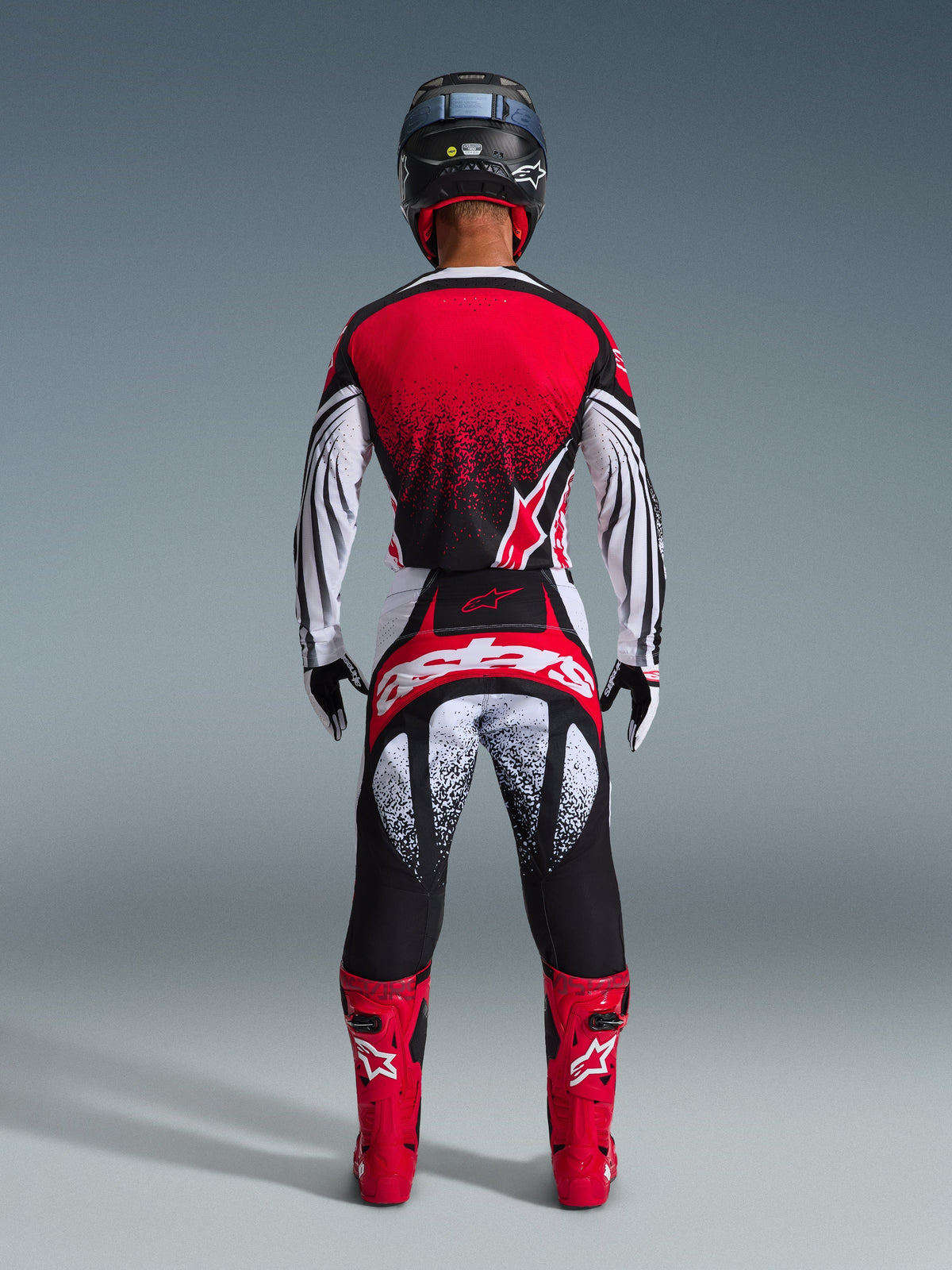 Pilote de motocross vu de dos portant le maillot Alpinestars Techstar Nomur 2026, rouge, noir et blanc avec un motif pointillé, pantalon de course assorti, bottes de motocross rouges et casque noir, tenue complète pour la compétition tout-terrain