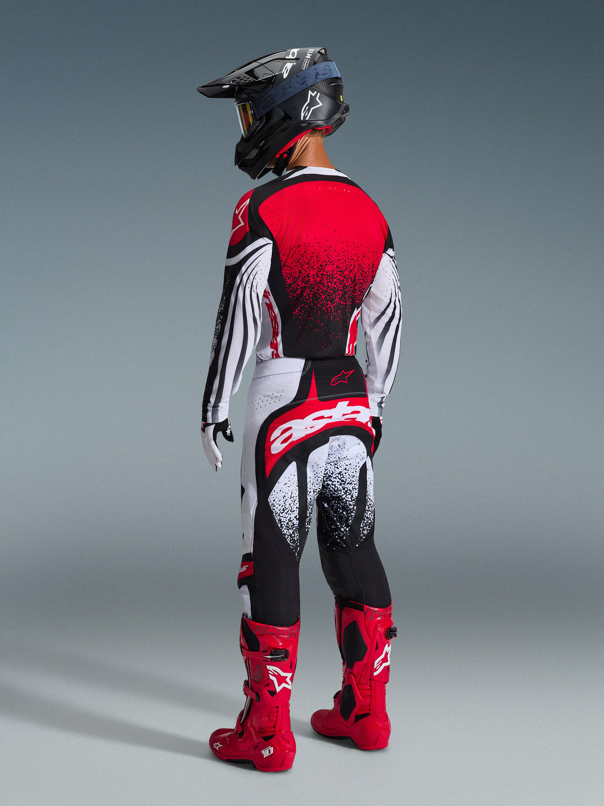 Pilote de motocross vu de dos portant l'ensemble d'équipement Alpinestars Techstar Nomur 2026, maillot rouge, blanc et noir avec dégradé à pois, pantalon assorti avec marquage 'astars' imposant, casque noir et bottes de motocross rouges, kit de course tout-terrain professionnel