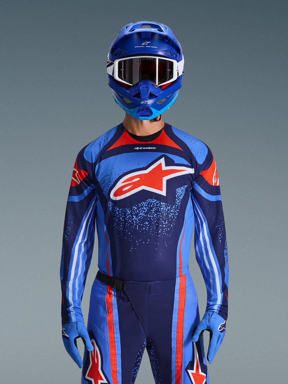 Maillot Alpinestars Techstar Nomur 2026, maillot de motocross, bleu marine foncé, orange et bleu UCLA, porté avec un pantalon assorti, un casque et des gants bleus, arborant un logo étoile blanche sur la poitrine avec des lignes dynamiques et des motifs mouchetés