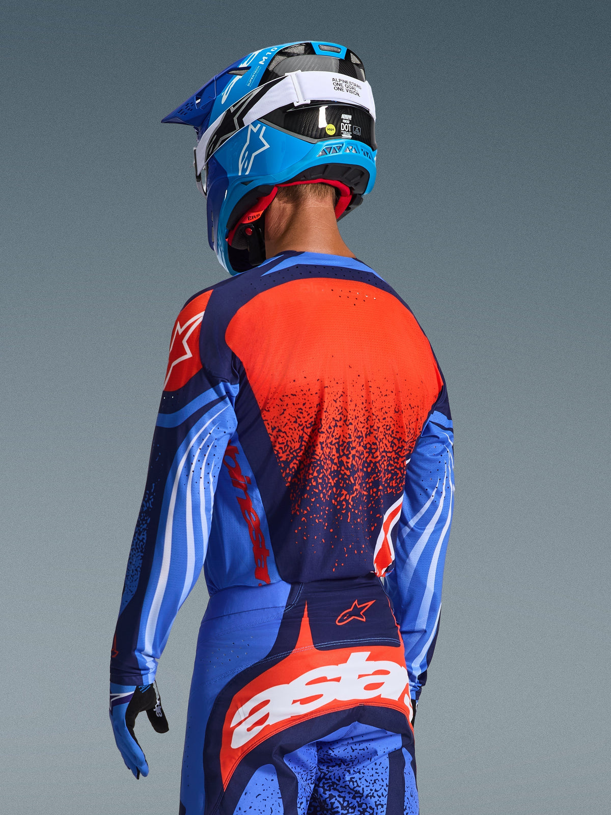 Pilote de motocross vu de dos portant le maillot Alpinestars Techstar Nomur 2026 et le pantalon assorti, bleu marine foncé et bleu UCLA avec des accents orange vifs, présentant un grand graphisme d'étoile et des ventilations découpées au laser sur le maillot, associé à un casque bleu et blanc pour une tenue de course tout-terrain complète.