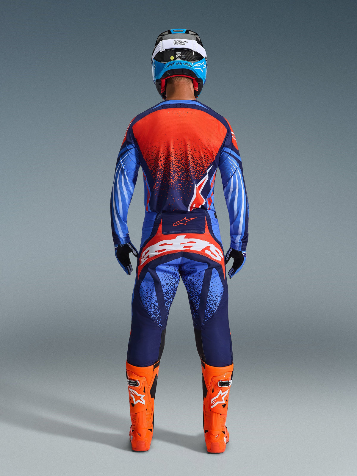 Pilote de motocross de dos portant l'ensemble Alpinestars 2026 Techstar Nomur en bleu marine foncé, orange et bleu UCLA, maillot avec dos orange moucheté, pantalon assorti avec marquage blanc imposant sur l'assise, bottes orange vif et casque bleu et blanc, tenue de course tout-terrain coordonnée.