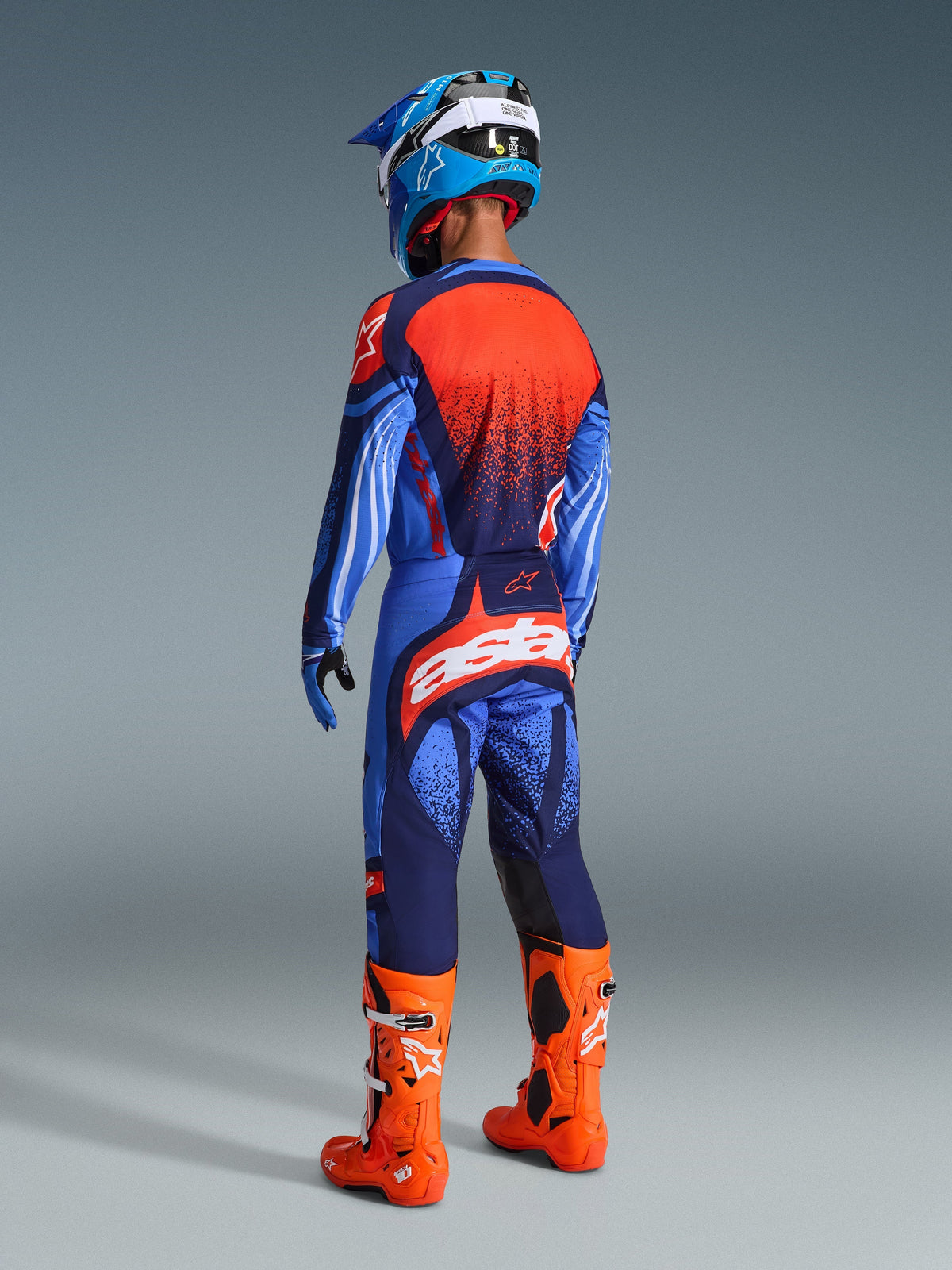 Pilote de motocross vu de dos portant le maillot Alpinestars Techstar Nomur 2026 en bleu marine foncé, orange et bleu UCLA, associé à un pantalon assorti avec logos blancs, bottes orange vif et casque bleu et blanc, équipement de course tout-terrain complet.