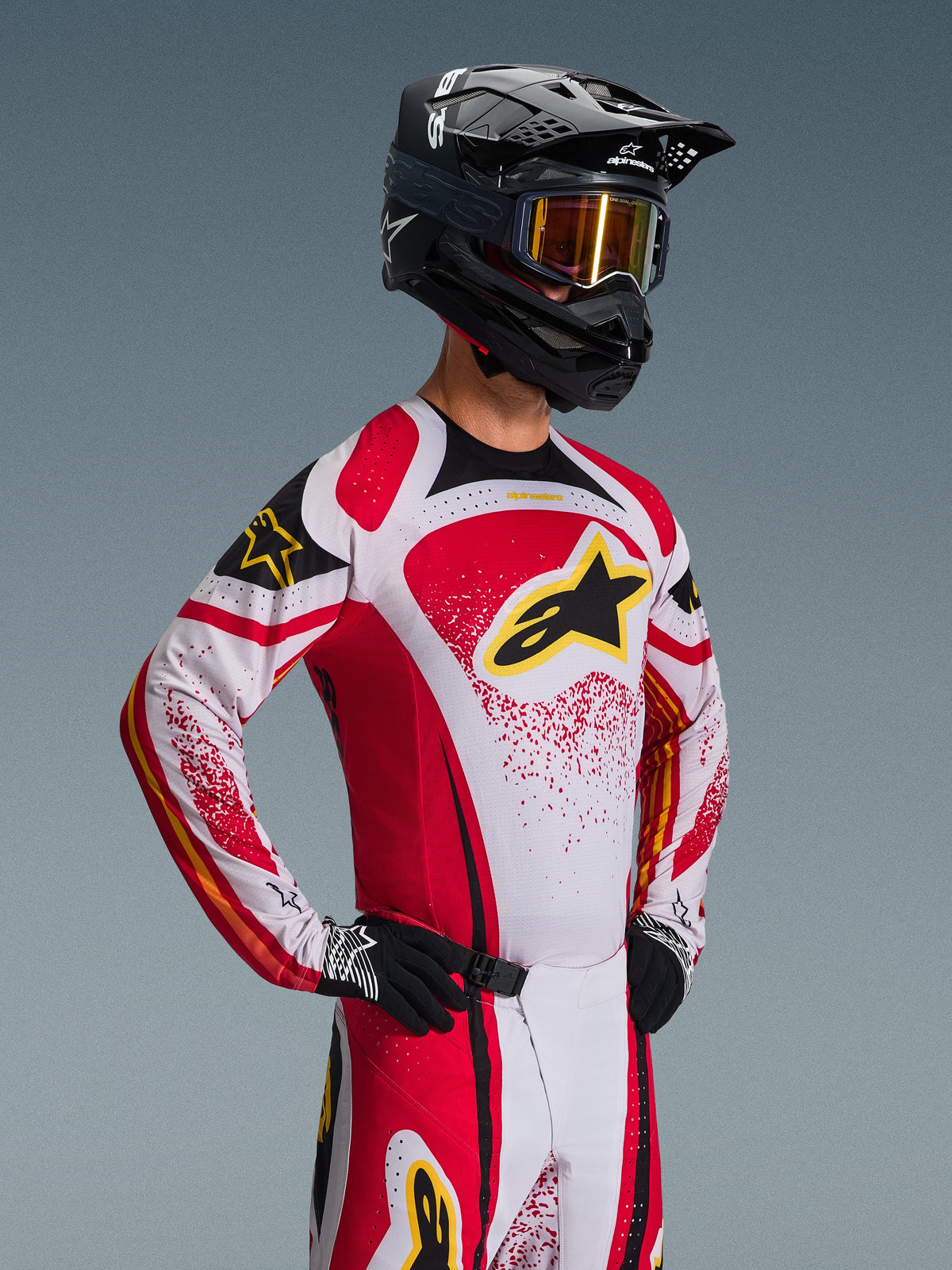 Pilote de motocross portant l'ensemble d'équipement Alpinestars 2026 Techstar Nomur, maillot blanc et rouge avec des accents noirs et un logo jaune, pantalon assorti, casque noir avec des lunettes teintées or, gants noirs et blancs, coupe de course athlétique pour la performance tout-terrain