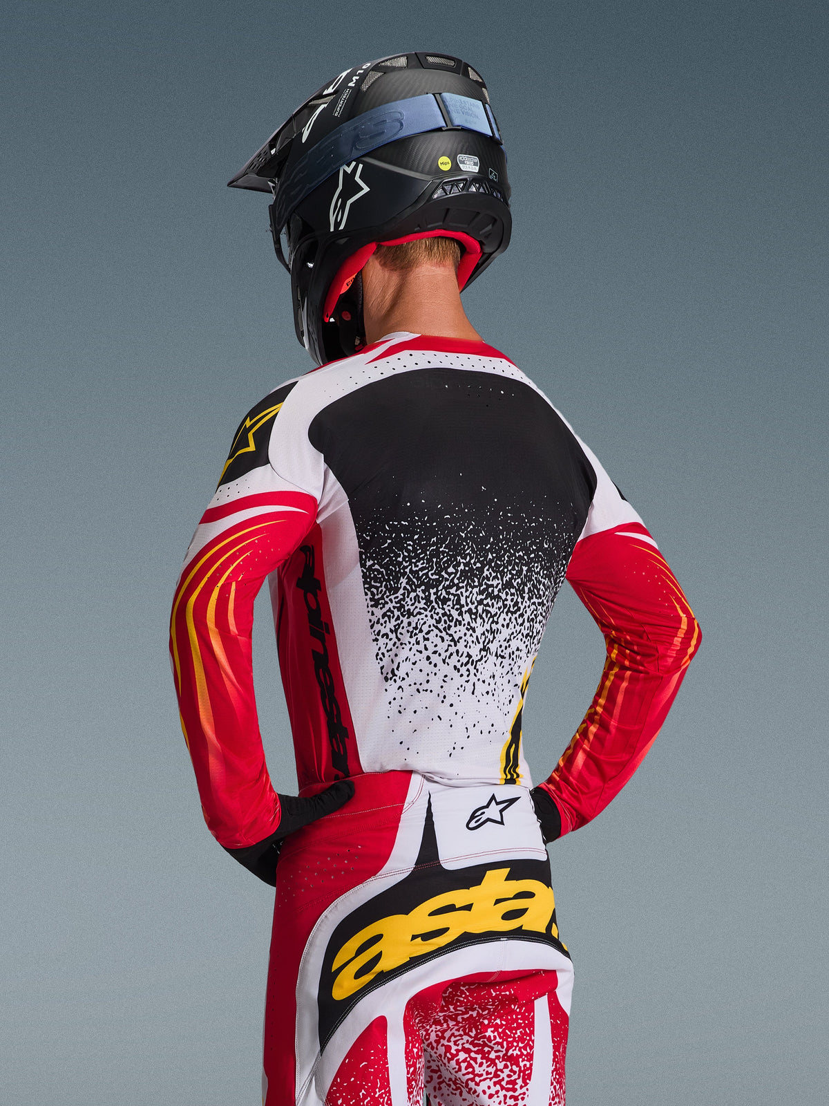 Ensemble d'équipement Alpinestars Techstar Nomur 2026, maillot et pantalon de motocross, rouge, blanc et noir avec des accents jaunes, vue arrière d'un pilote portant un casque noir, vêtements de course haute performance avec motifs mouchetés et logos audacieux