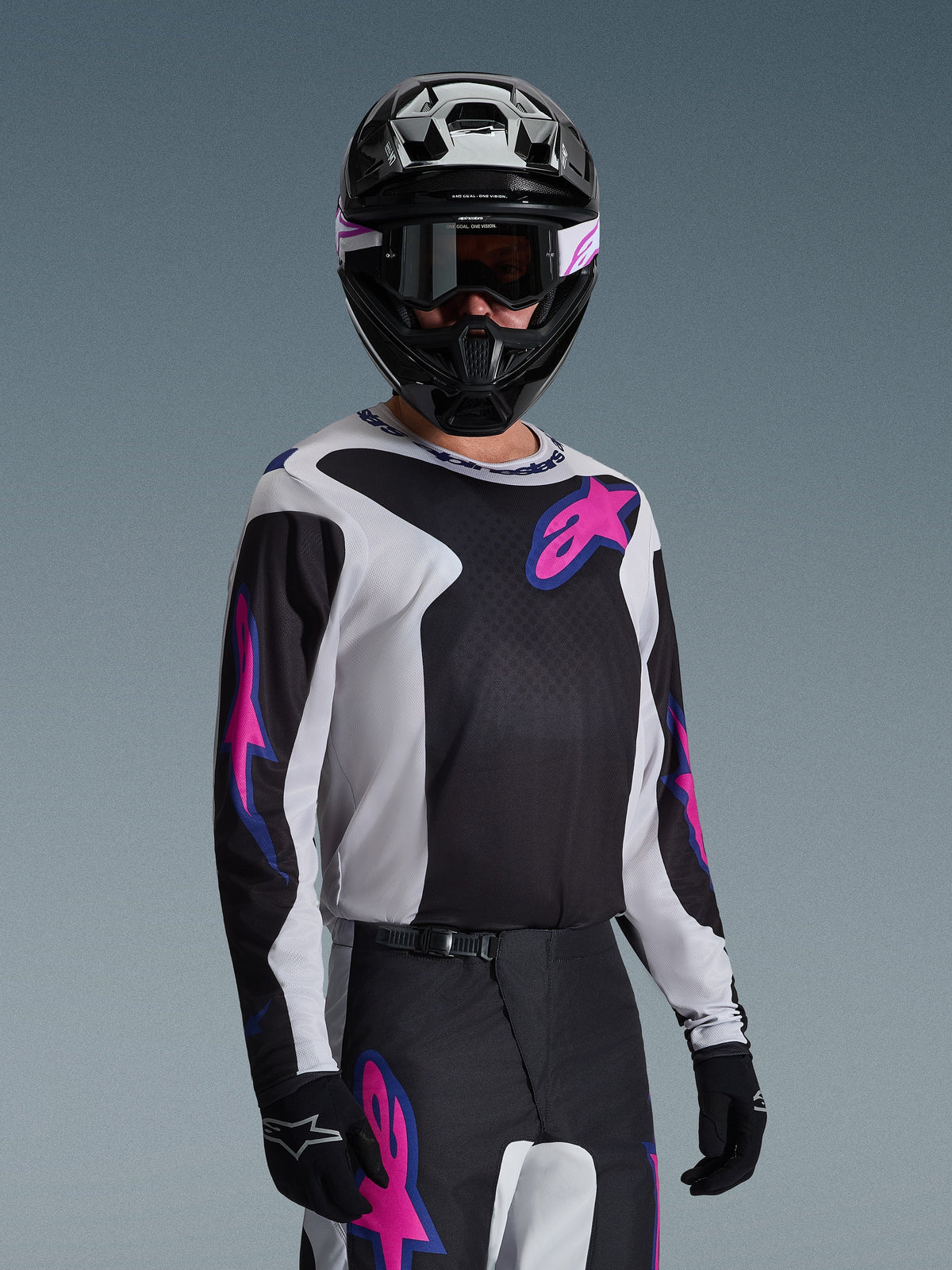 Motocross rider wearing the Alpinestars 2026 Fluid Grid Jersey, Black, Light Gray, and Purple, featuring large pink and purple star logos, paired with matching pants, black helmet, and goggles, complete off-road racing gear set
