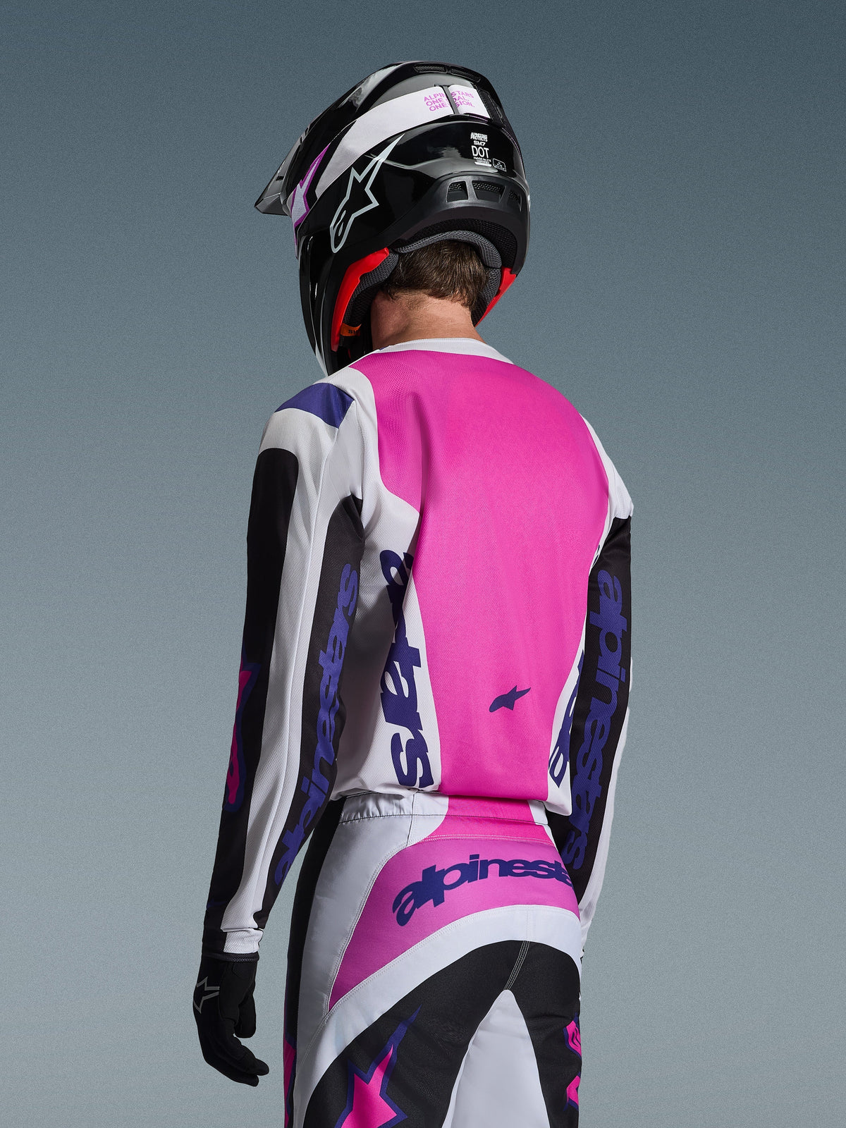 Pilote de motocross vu de dos portant le maillot Alpinestars 2026 Fluid Grid en noir, blanc et violet, pantalon assorti avec marquage audacieux, casque noir avec des touches de rose et gants noirs, tenue coordonnée complète pour la performance tout-terrain.