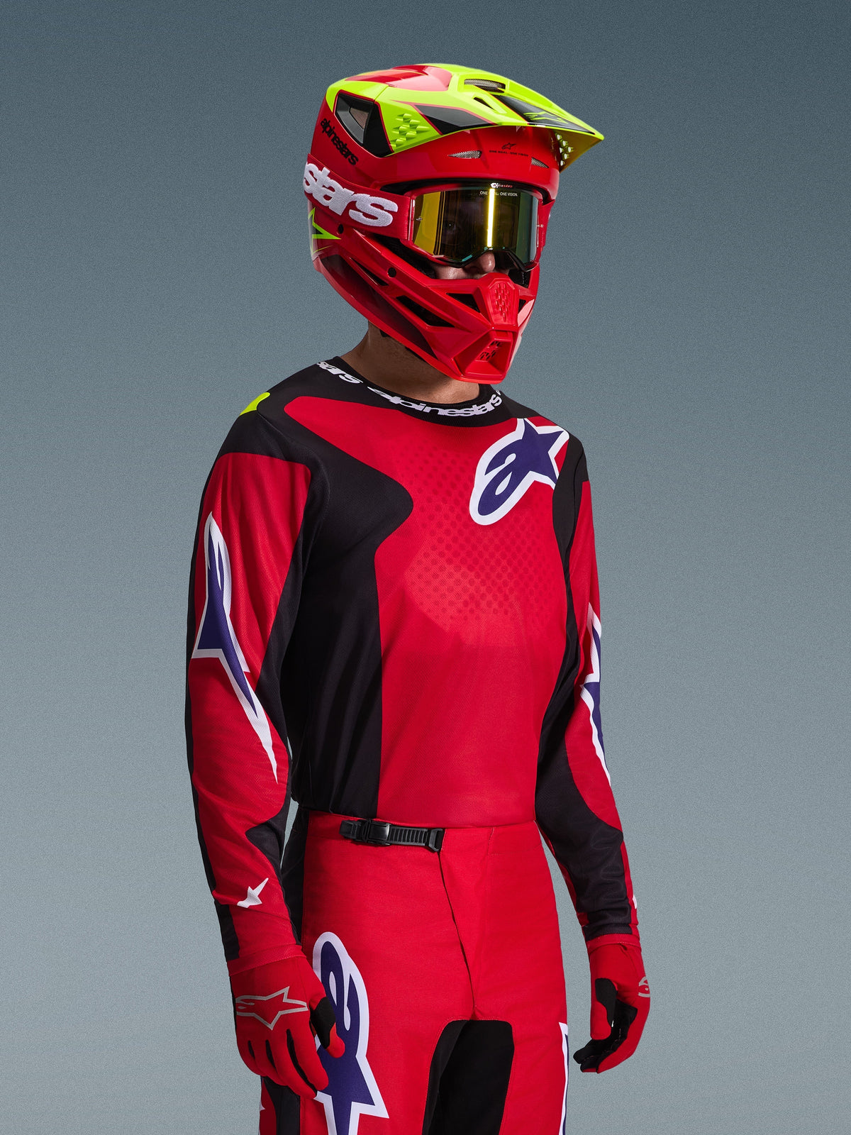 Motocross rider wearing the Alpinestars 2026 Fluid Grid Jersey in Red Black and Purple, coordinated with matching motocross pants, a red helmet with neon yellow accents, gold goggles, and red gloves, complete off-road racing outfit.