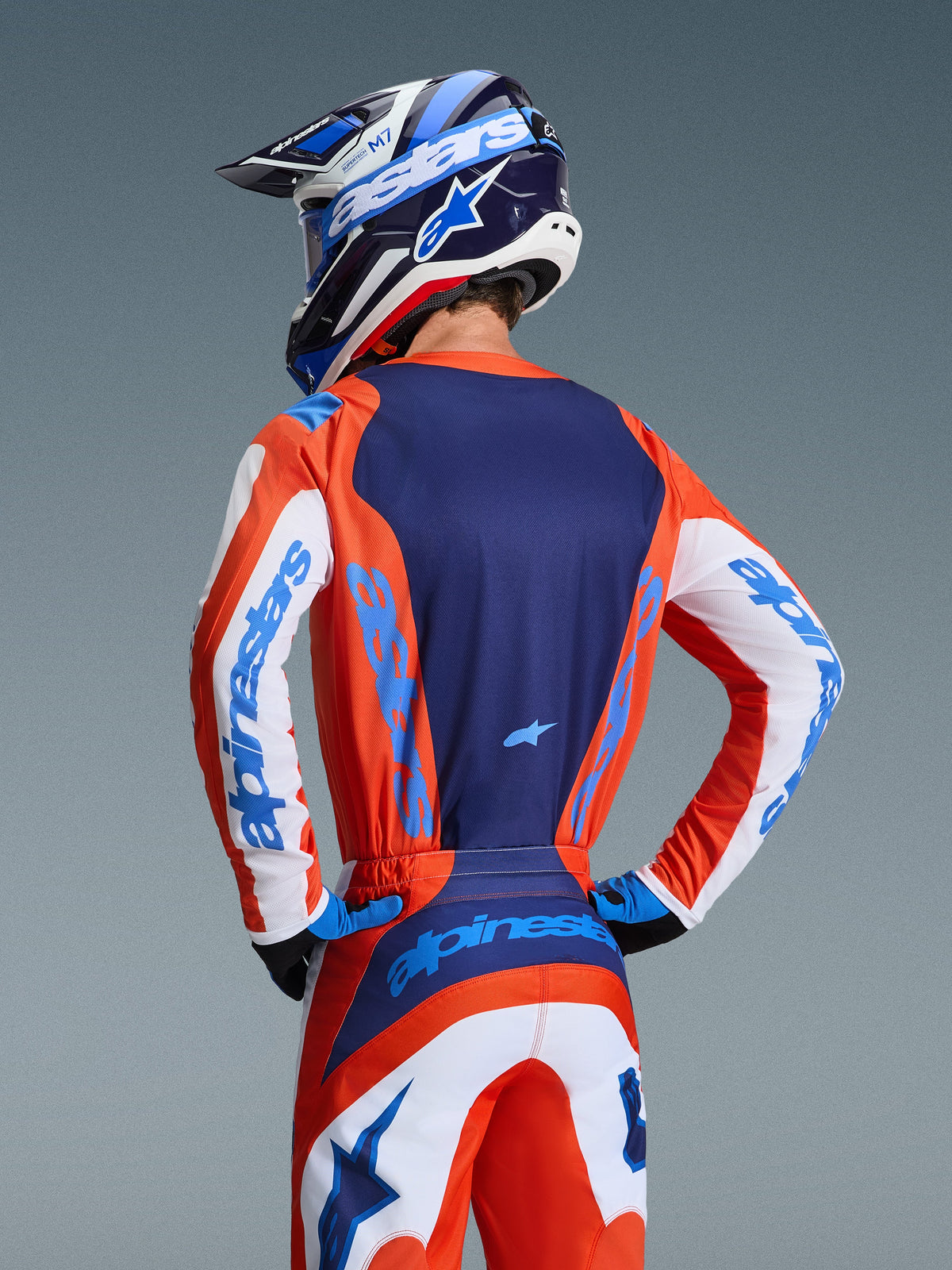 Maillot Alpinestars Fluid Grid 2026, maillot de motocross, orange et bleu UCLA, vue arrière d'un pilote portant le pantalon et le casque assortis, avec panneau arrière bleu marine et manches blanches avec logos bleu clair, coupe athlétique pour le tout-terrain